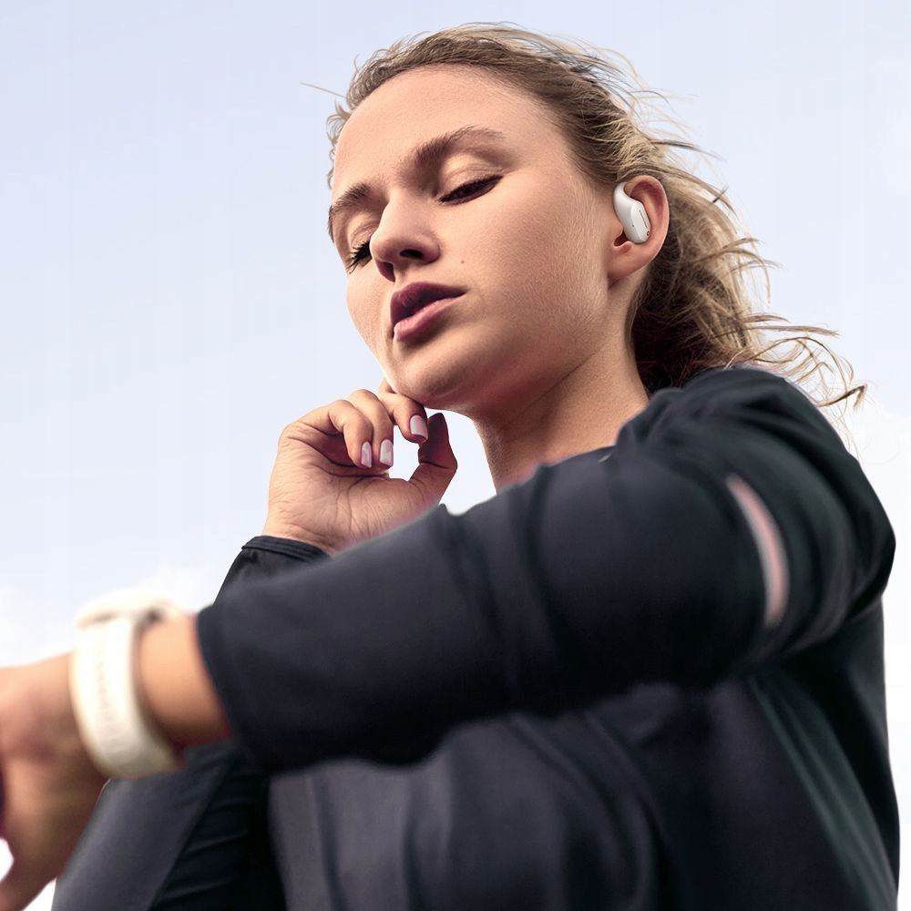 Una donna in abbigliamento sportivo e auricolari bianchi guarda il suo smartwatch sotto un cielo azzurro.