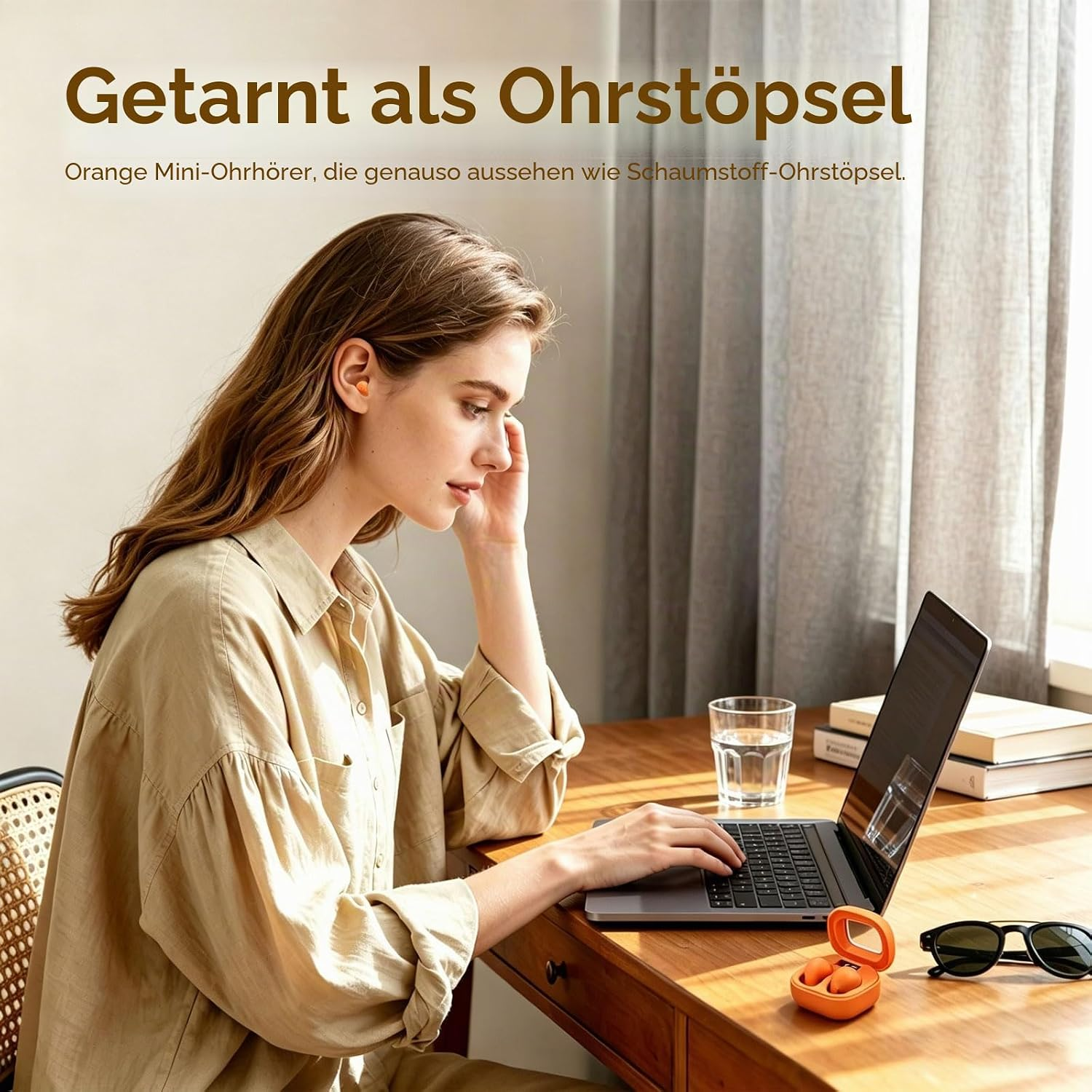 Eine blonde Frau arbeitet an einem Laptop an einem Holztisch, mit kleinen orangefarbenen Ohrhörern im Ohr.