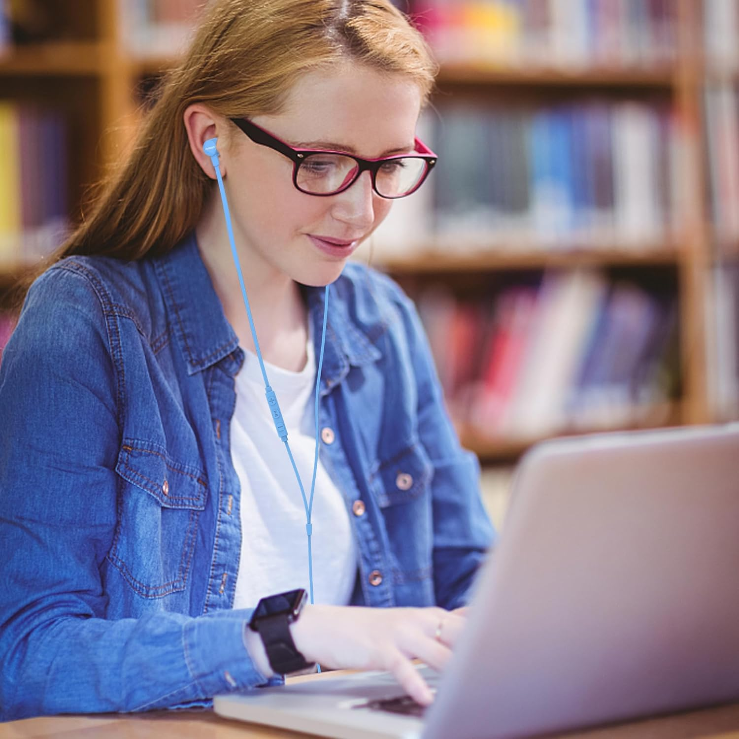 Eine junge Frau mit rotem Haar und Brille tippt in einer Bibliothek an einem Laptop und trägt blaue Kopfhörer.