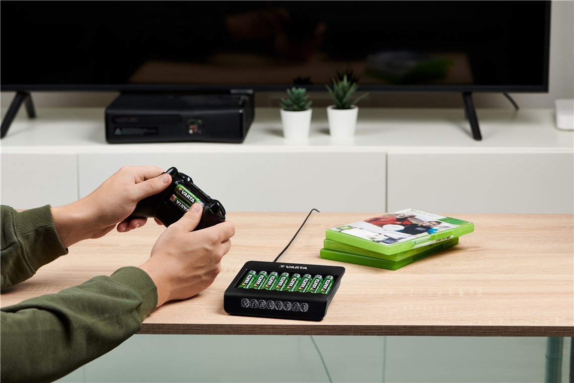 Persona inserta pilas Varta en un mando de Xbox, con un cargador y videojuegos cerca.