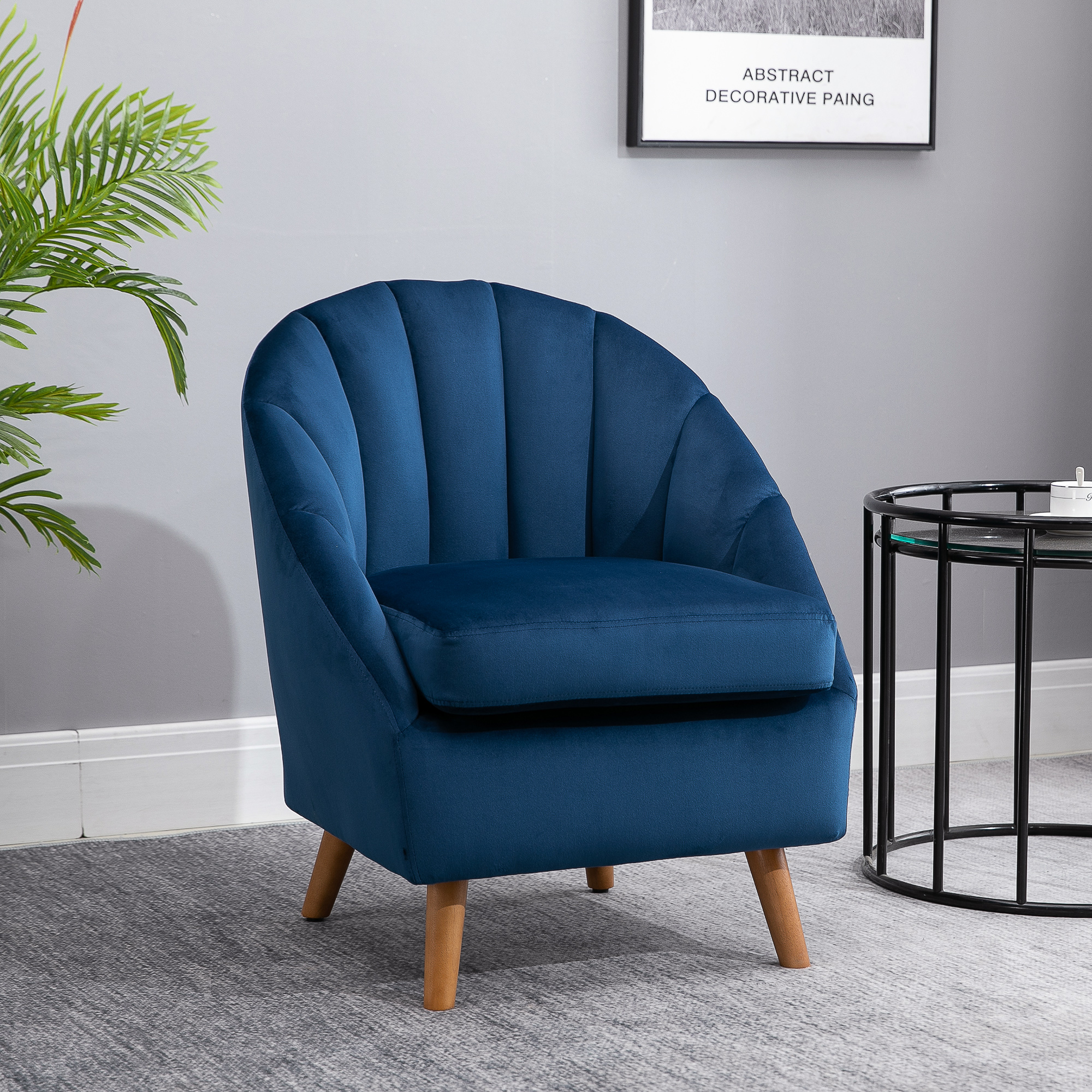 Poltrona in velluto blu con gambe in legno contro una parete grigia, accanto a un tavolino e a una pianta.