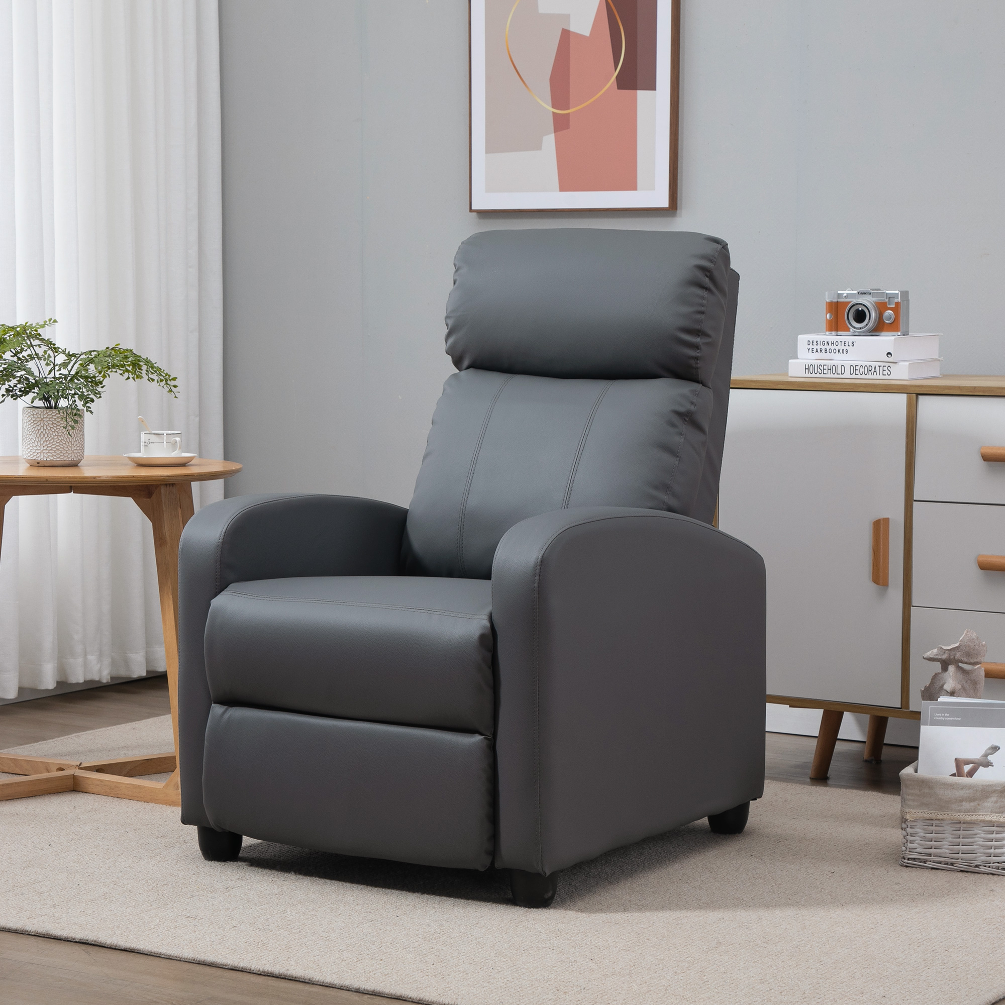 Una poltrona in pelle grigia si trova su un tappeto beige in una stanza con tavolino, mobile, pianta e quadro.