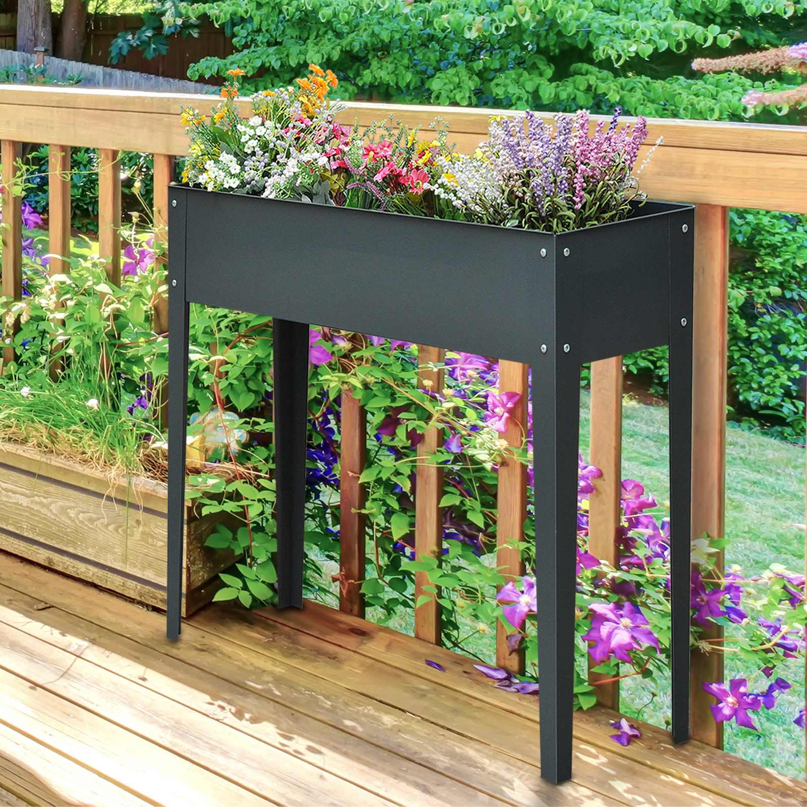 Una fioriera rialzata grigia con gambe, piena di fiori colorati, si trova su una terrazza in legno davanti a una ringhiera.