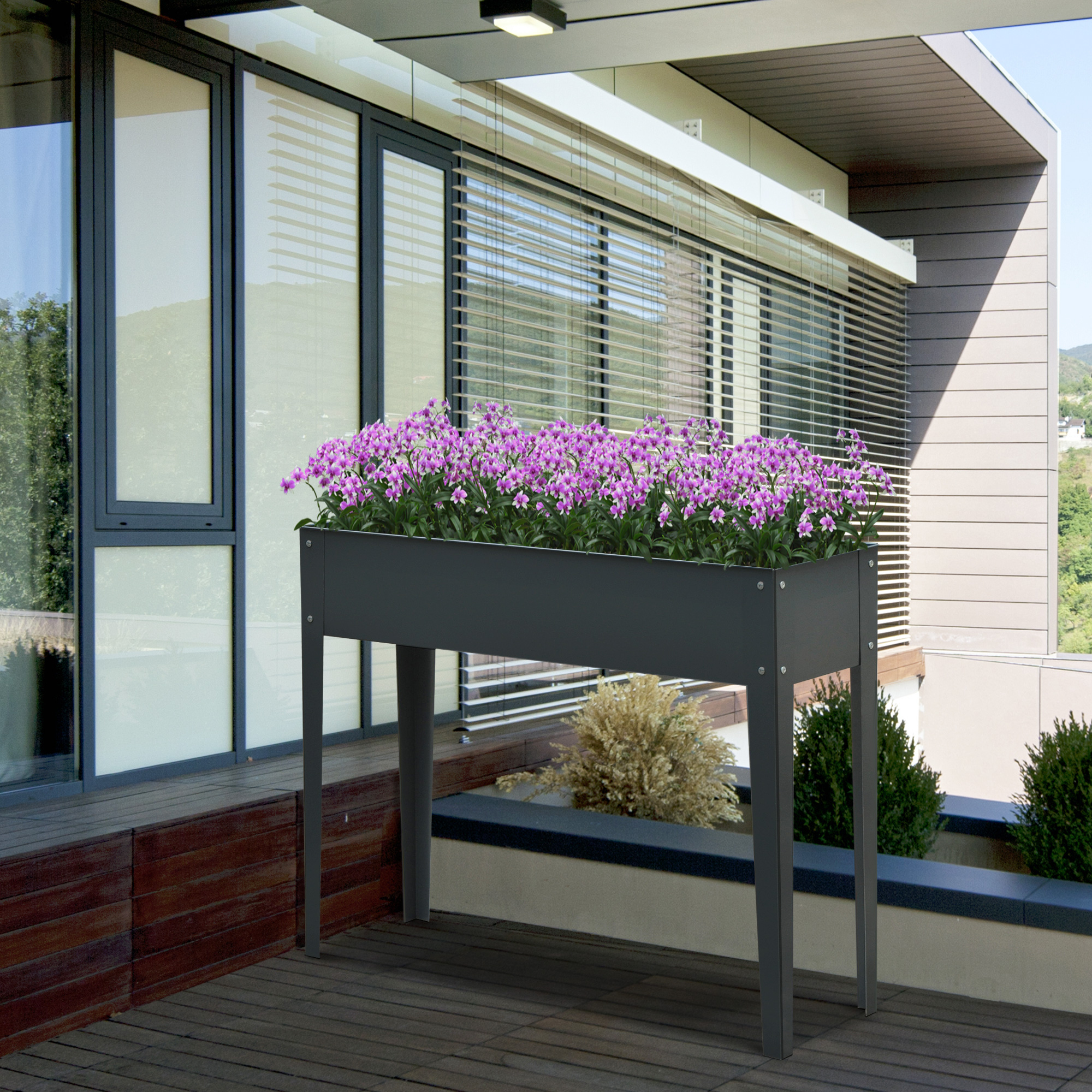 Una fioriera rialzata grigia con fiori viola si trova su una terrazza in legno davanti a un edificio moderno con persiane.
