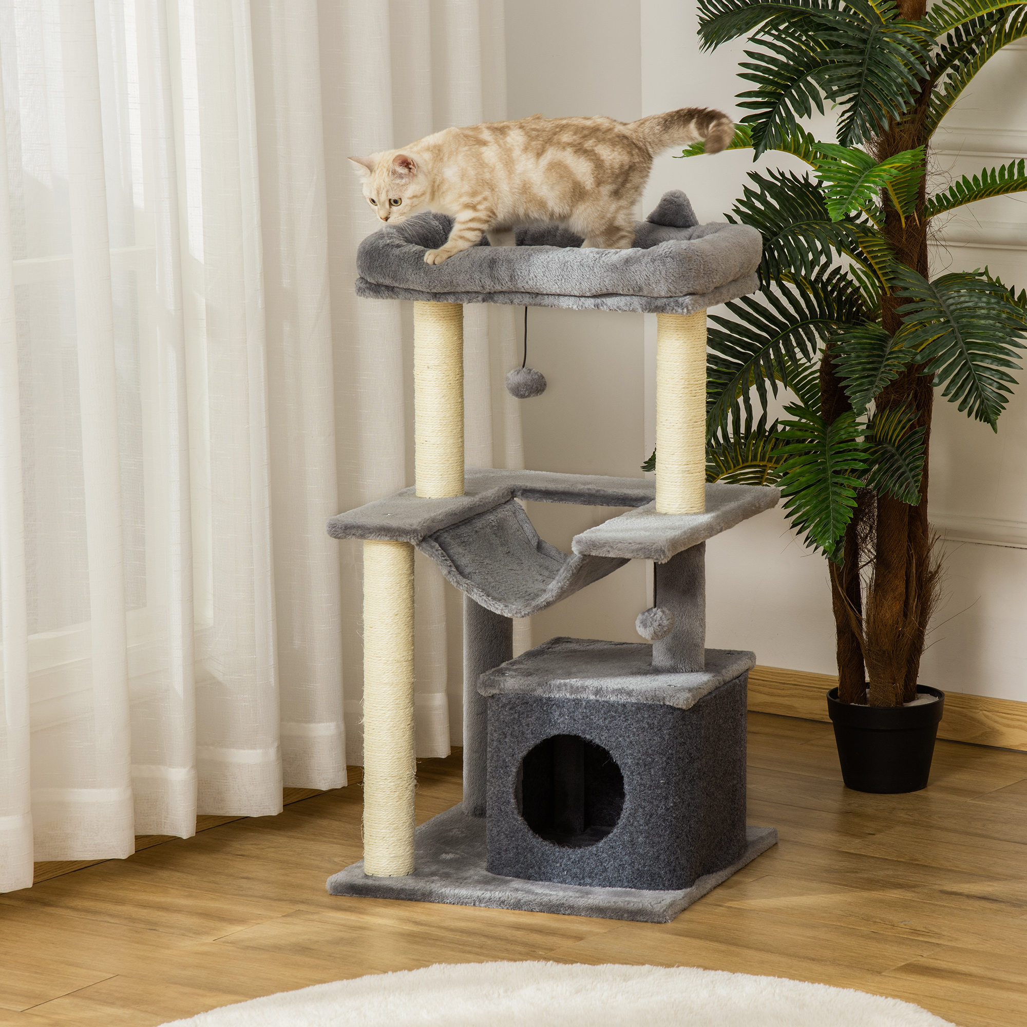 Un gatto tabby marrone chiaro si trova sulla piattaforma superiore di un tiragraffi grigio vicino a una pianta verde.