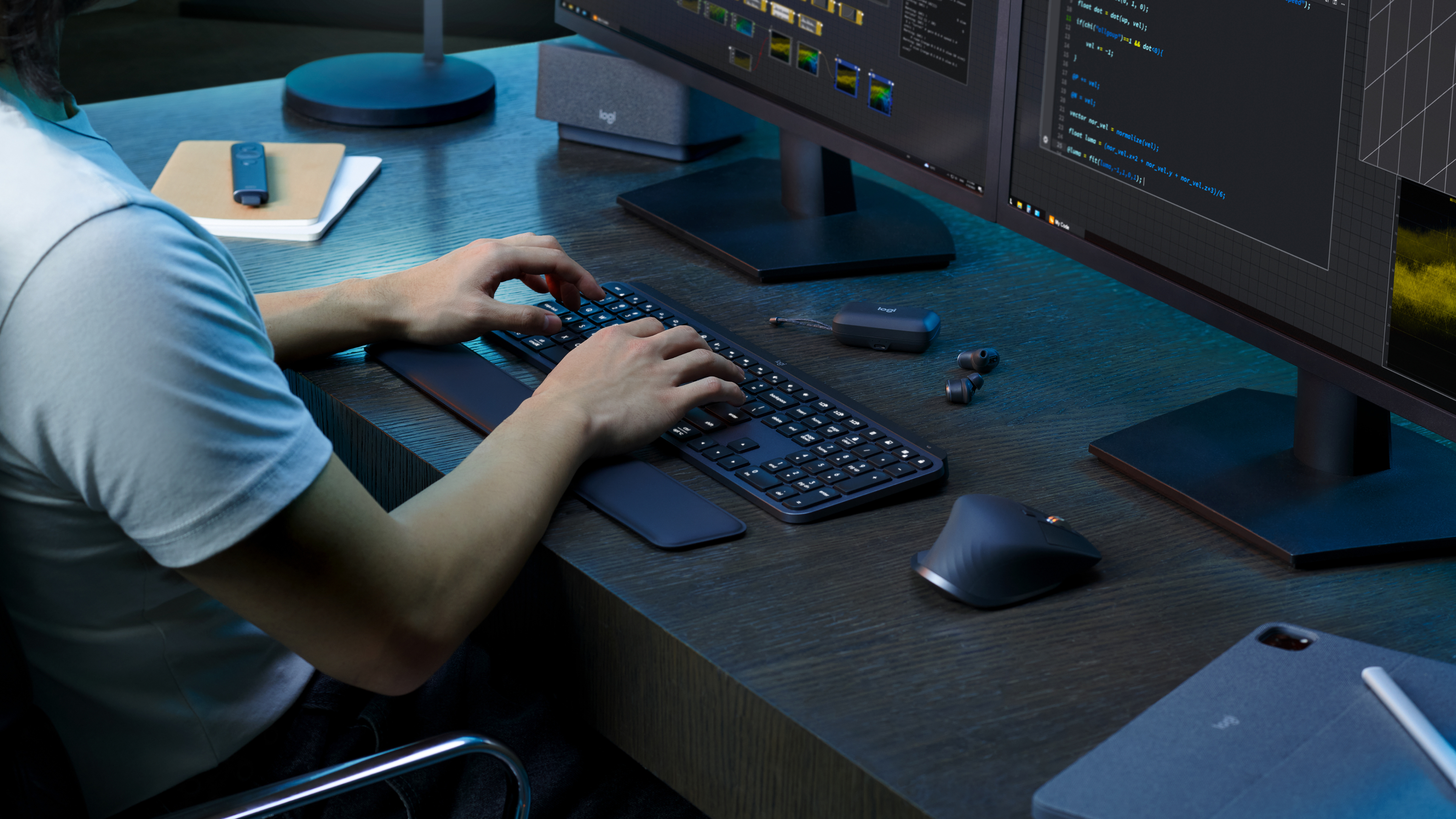 Persona che lavora a una scrivania in legno con tastiera, mouse e due grandi monitor sotto luce blu.