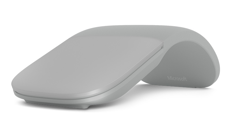 Un ratón Microsoft Arc gris reposa sobre una superficie blanca con el logo de Microsoft visible.