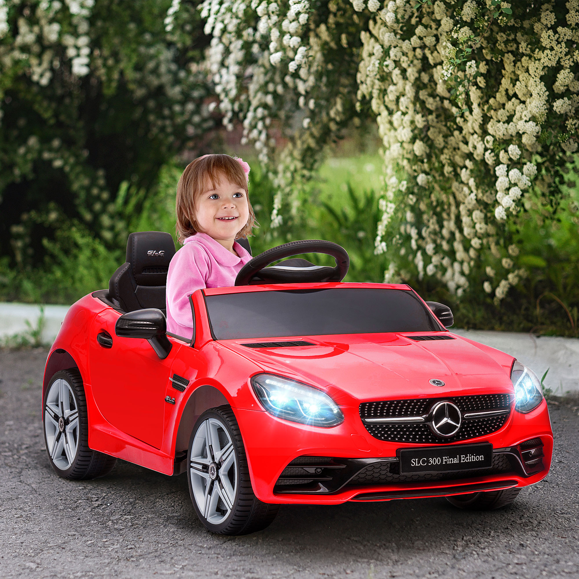 Una bambina siede su una macchinina rossa davanti a fiori bianchi.