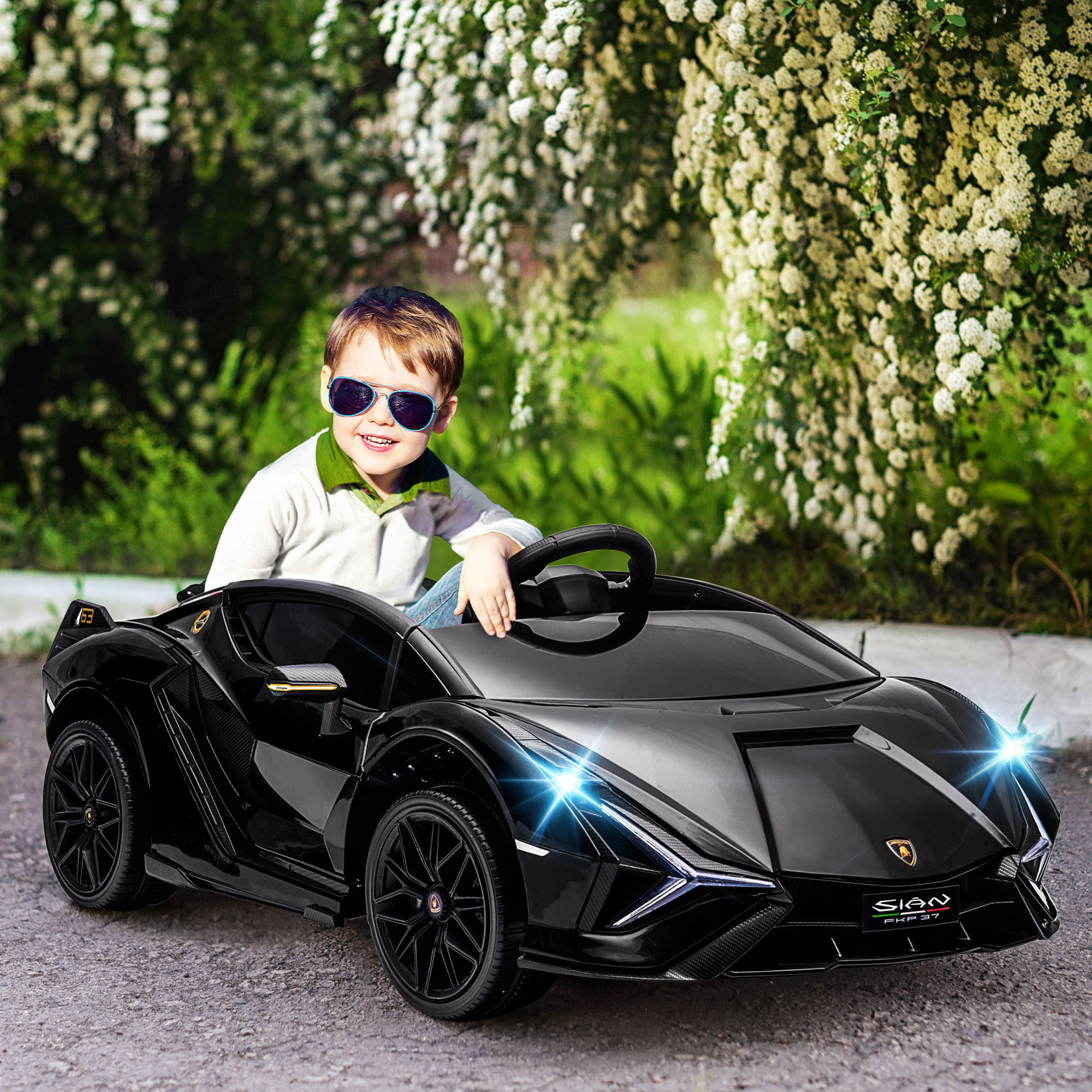 Un ragazzo con gli occhiali da sole siede sorridente in un'auto giocattolo nera davanti a cespugli fioriti bianchi.