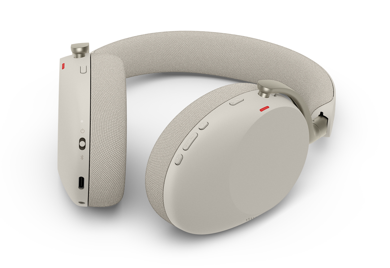 Cuffie over-ear beige su fondo bianco, con archetto in tessuto, pulsanti di controllo e porta di ricarica.