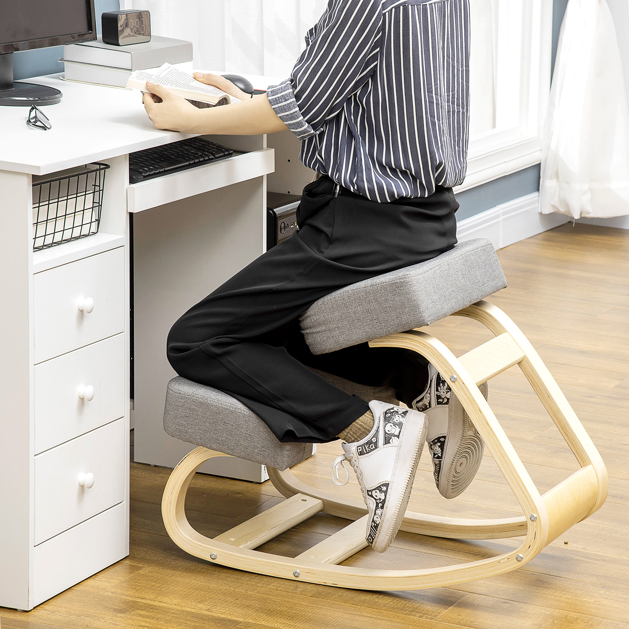 Una persona siede su una sedia ergonomica in legno per ginocchia a una scrivania bianca, leggendo un libro.