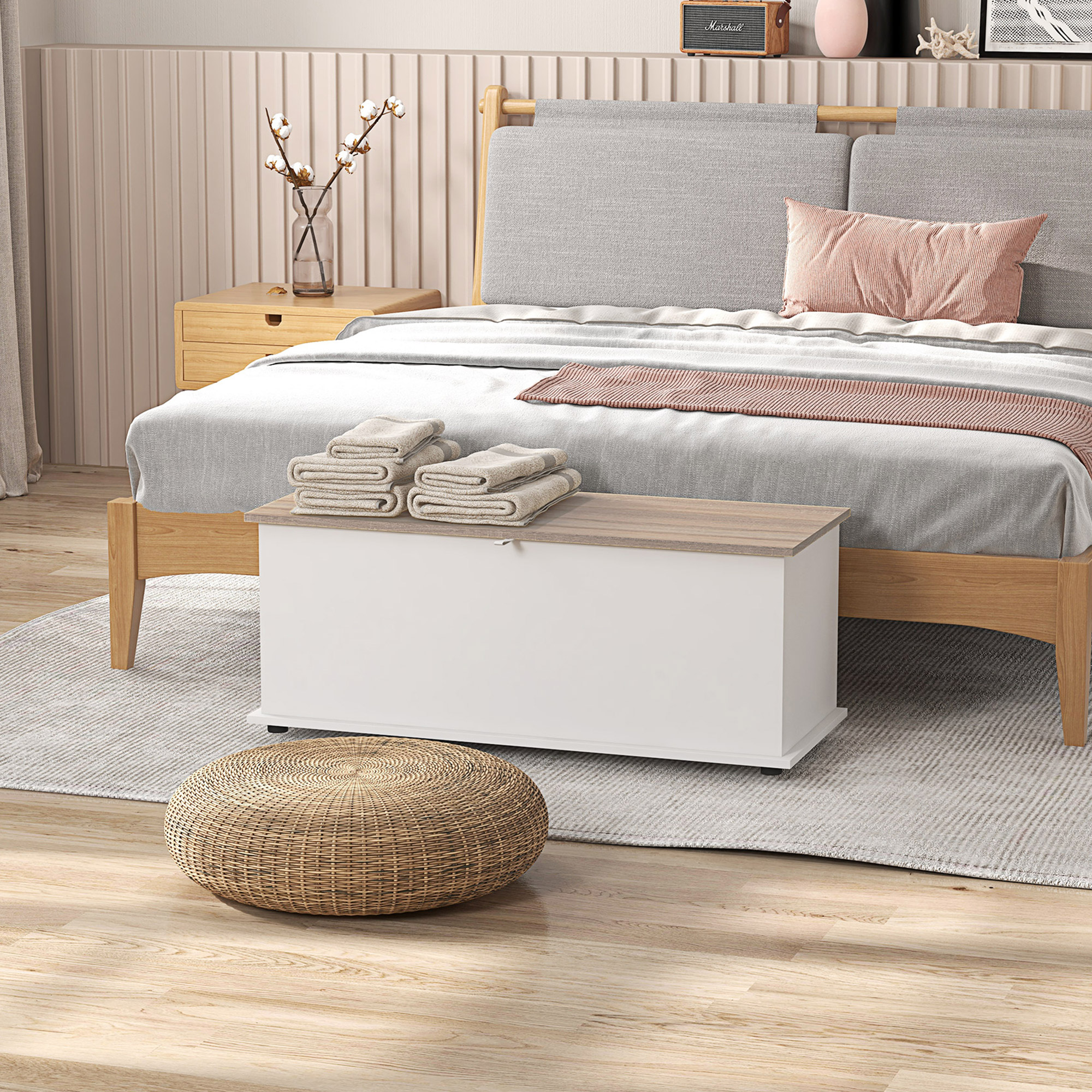 Camera da letto con letto in legno, biancheria grigia, panca contenitore bianca, asciugamani piegati e pouf intrecciato.