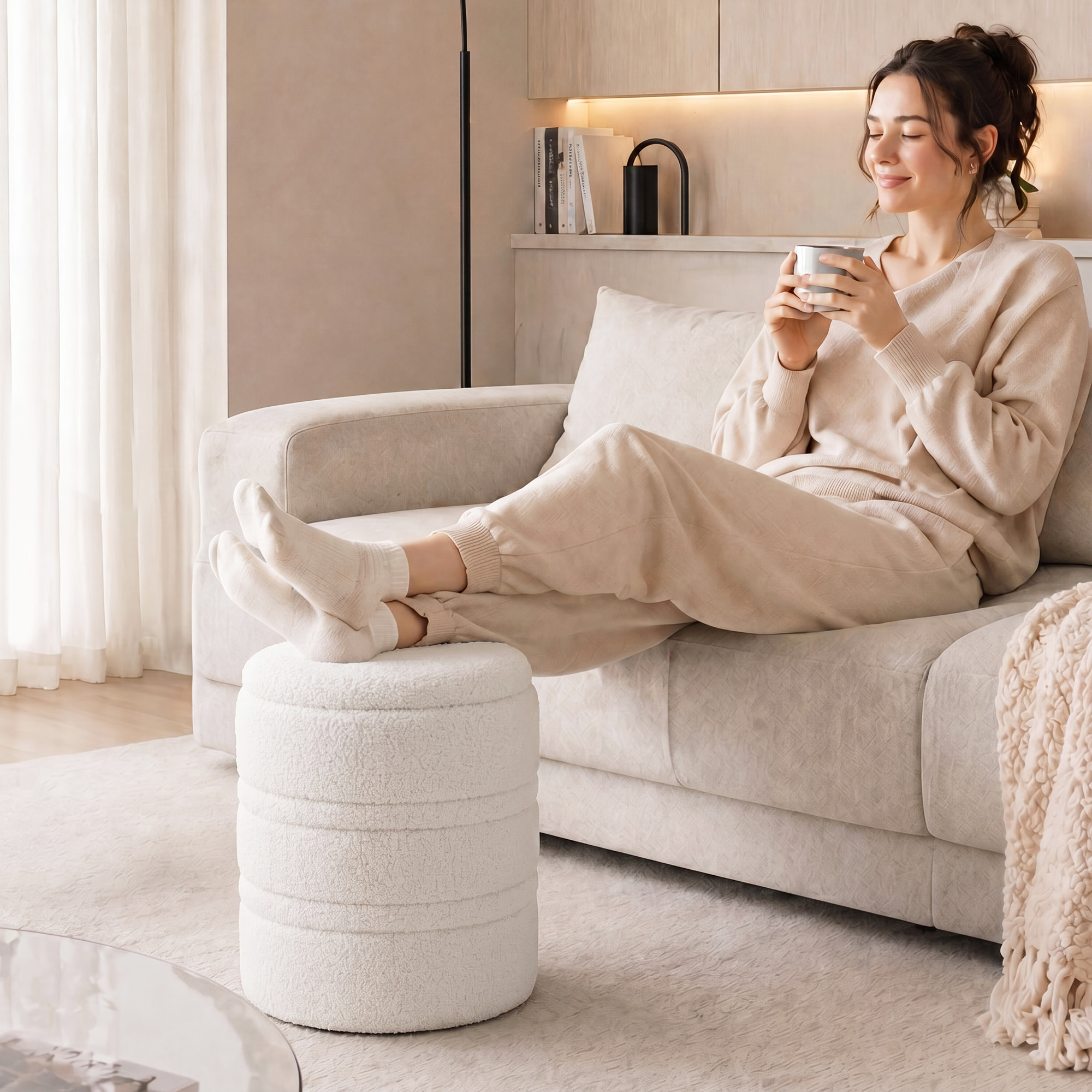 Una donna in abiti beige si rilassa su un divano, tenendo una tazza con i piedi su un pouf.