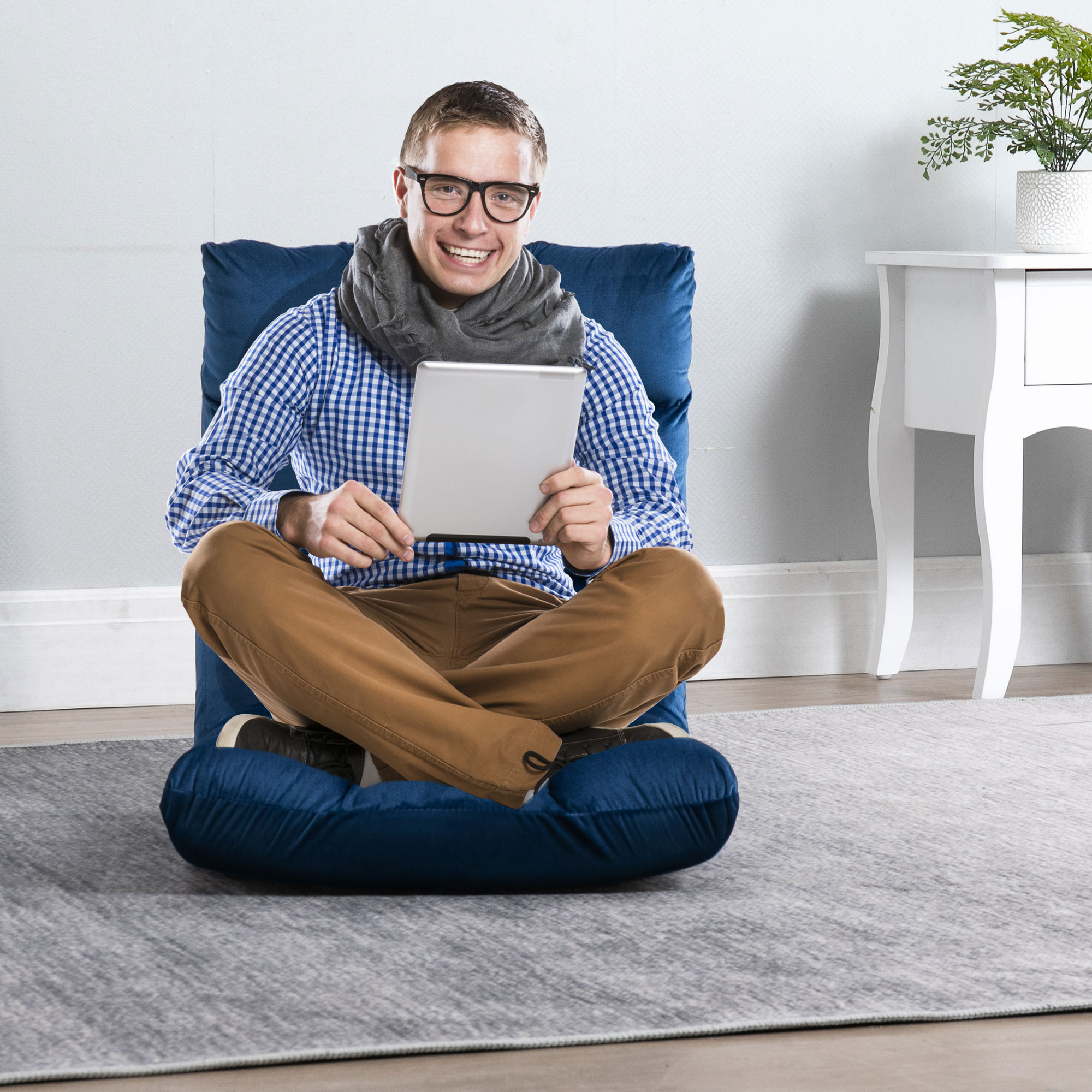 Un uomo sorridente con gli occhiali siede su un cuscino da pavimento blu, tenendo un tablet bianco.