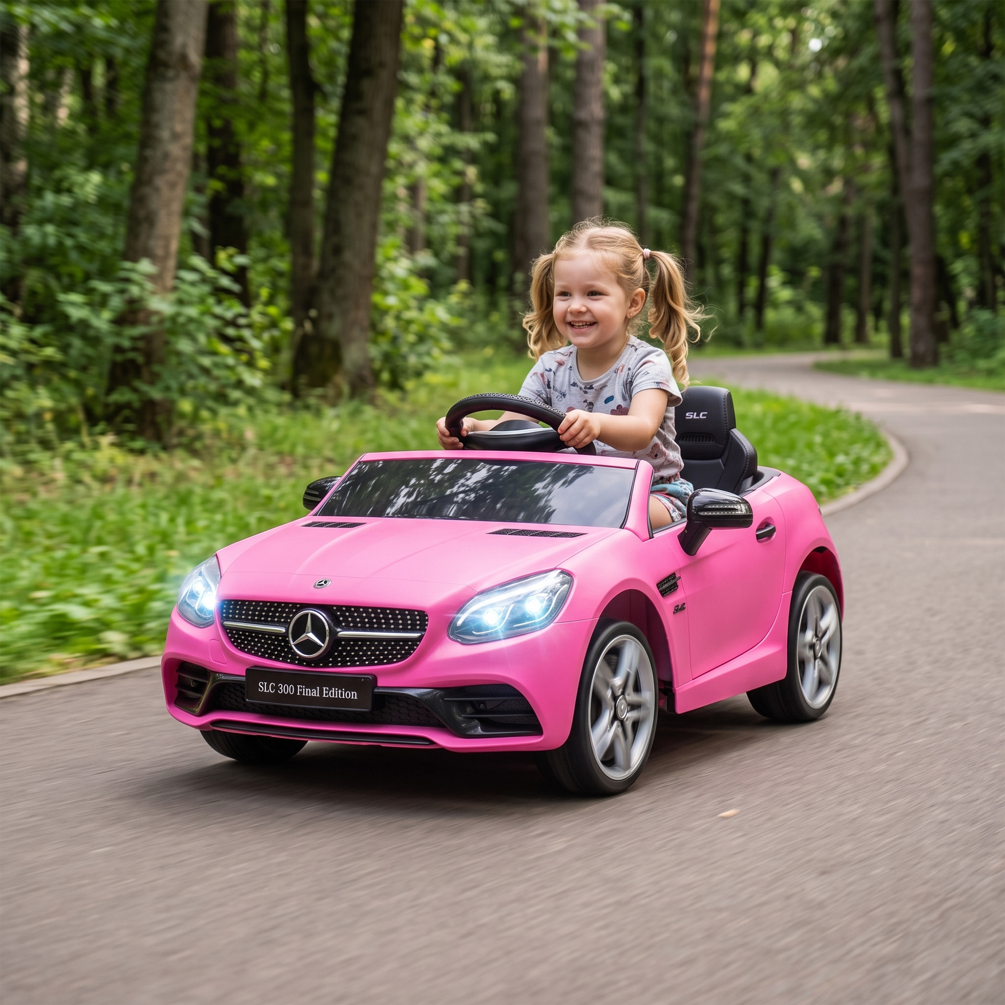 Una bambina sorridente con le codine guida un'auto giocattolo rosa su un sentiero asfaltato nel bosco.