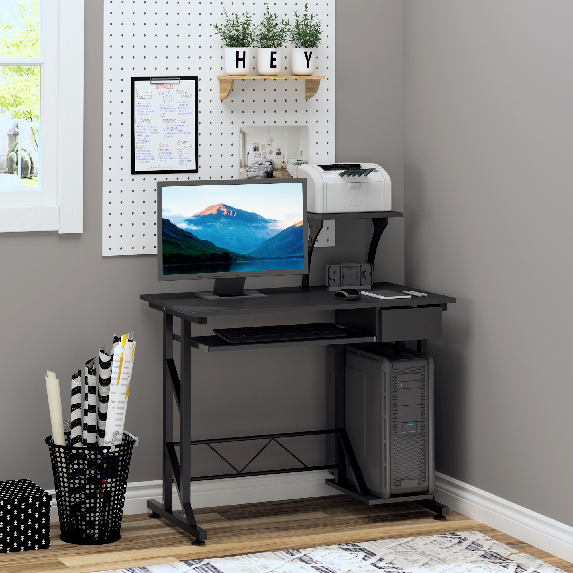 Una scrivania nera con monitor, PC, stampante e note su un pannello forato bianco.