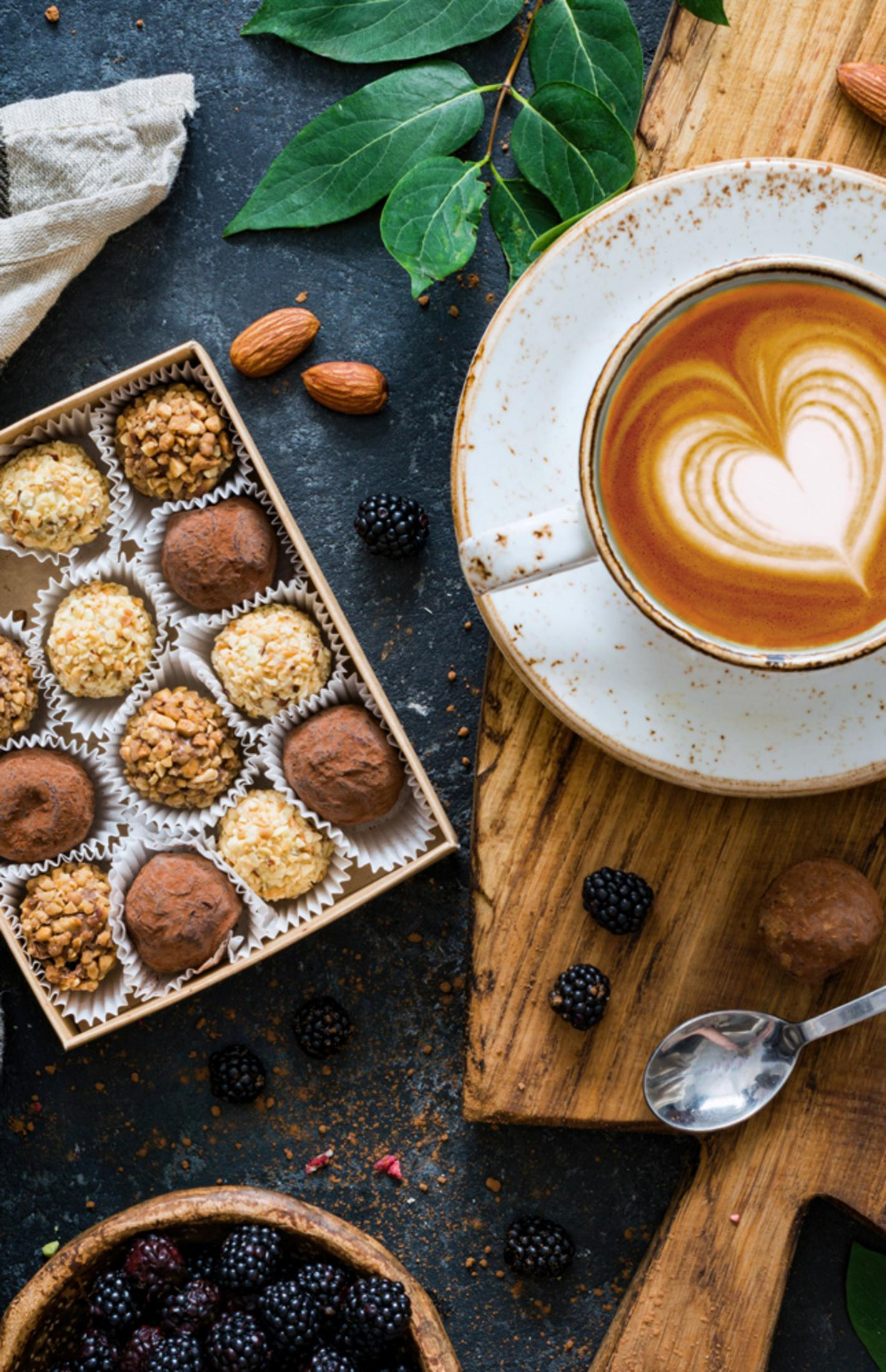 Eine Tasse Cappuccino mit Herzmuster, Pralinenschachtel, Brombeeren, Mandeln und ein Löffel auf Holzbrett.