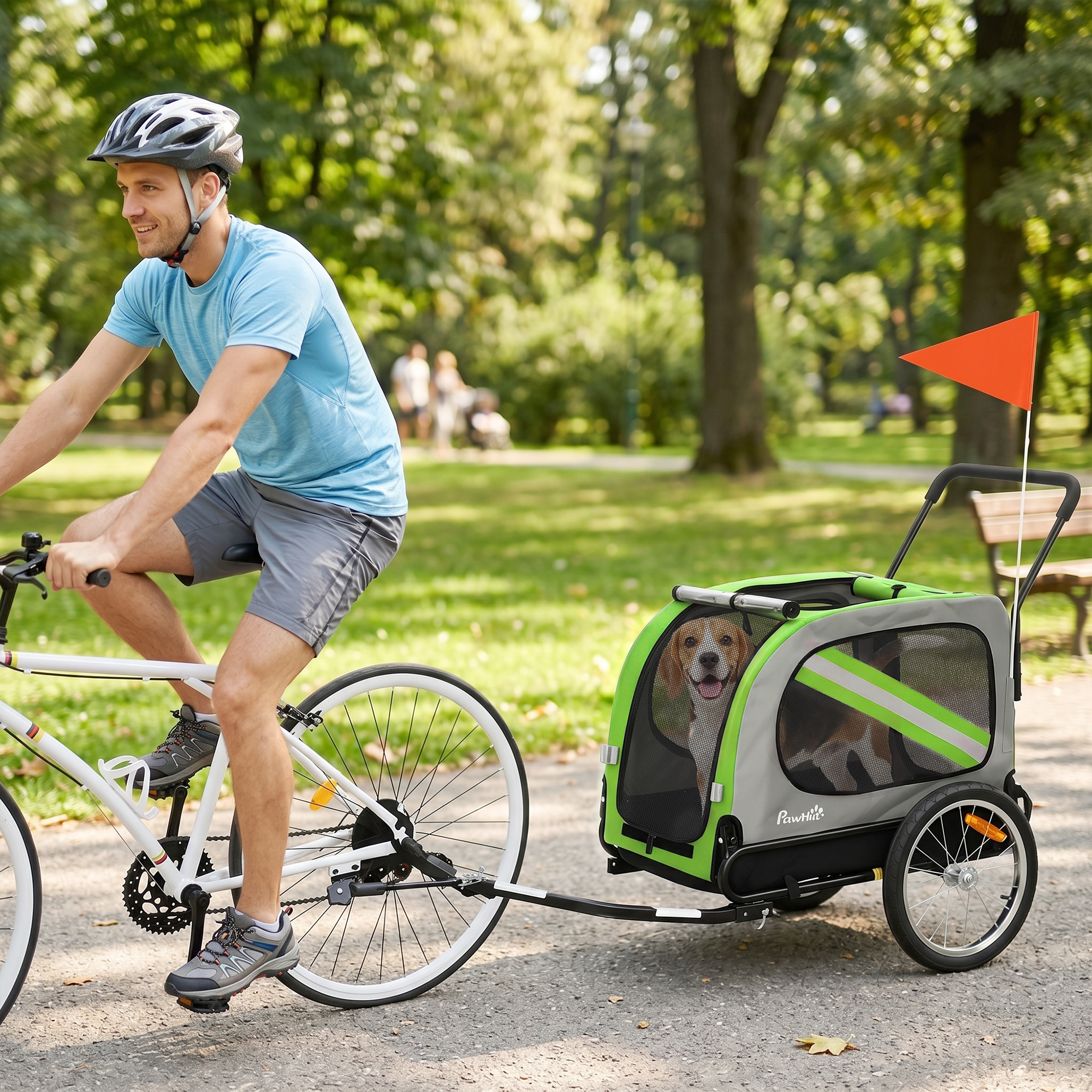 Un hombre con camisa azul monta en bicicleta, remolcando un carrito verde y gris con un perro dentro en un parque.