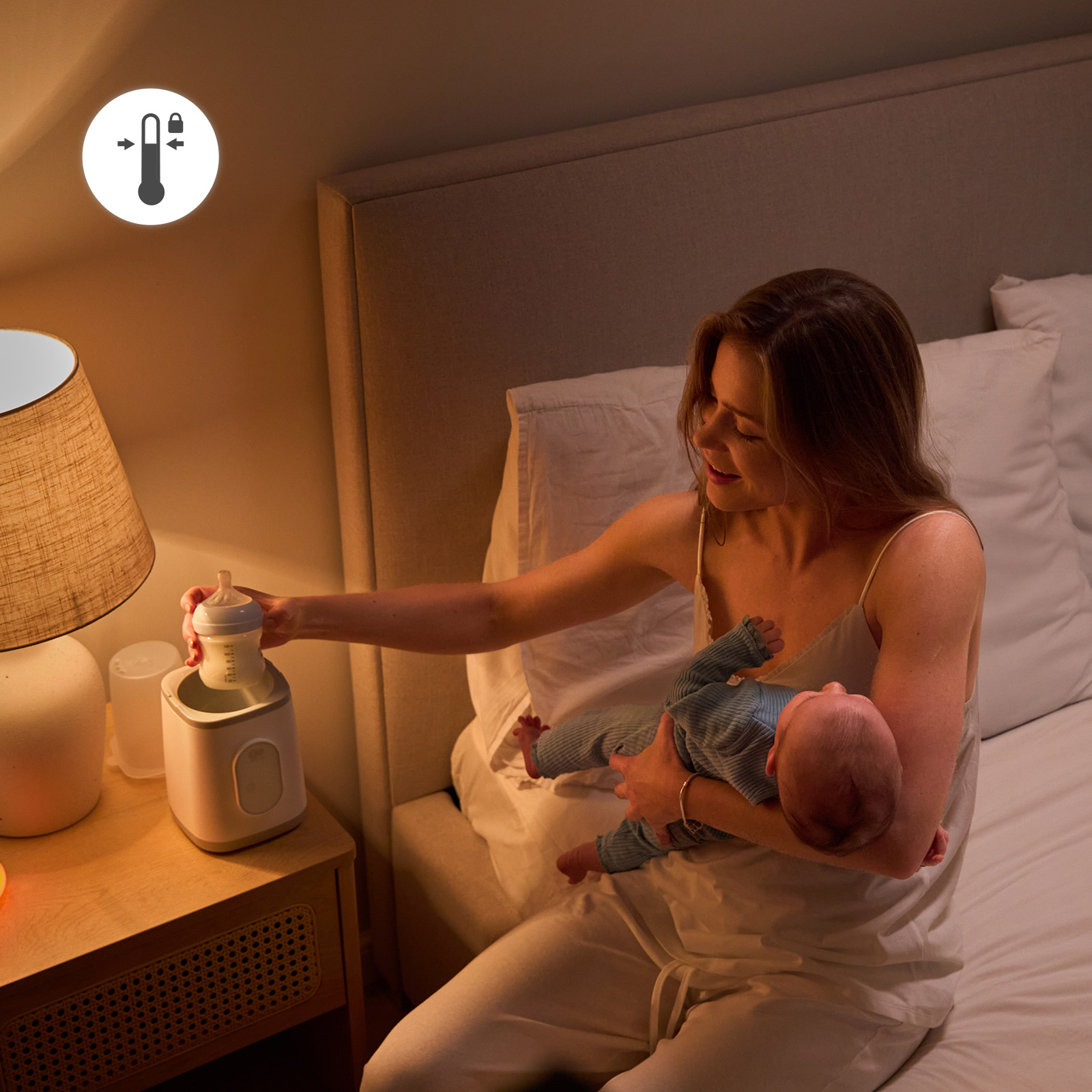 Eine Mutter sitzt im Bett, hält ein Baby und stellt eine Flasche in einen Flaschenwärmer auf den Nachtisch.