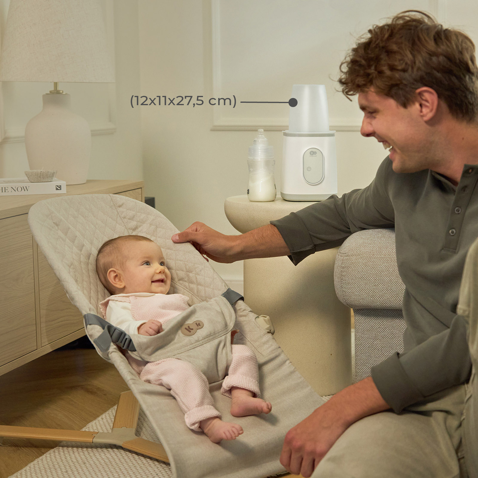 Ein Vater lächelt sein glückliches Baby in einer grauen Babyliege an, neben einem Flaschenwärmer auf einem Tisch.