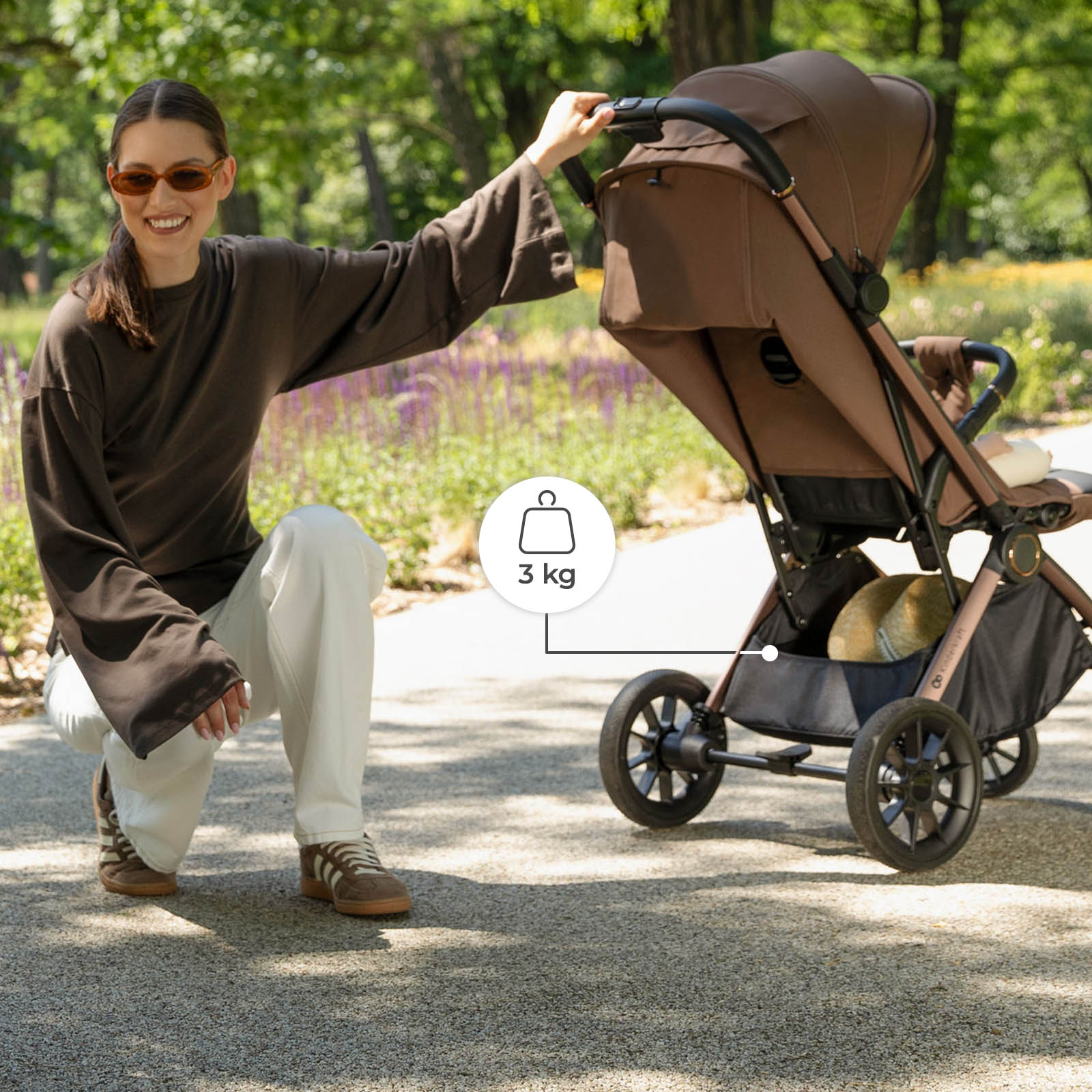 Eine Frau hockt neben einem braunen Kinderwagen auf einem Parkweg und zeigt auf den Einkaufskorb.