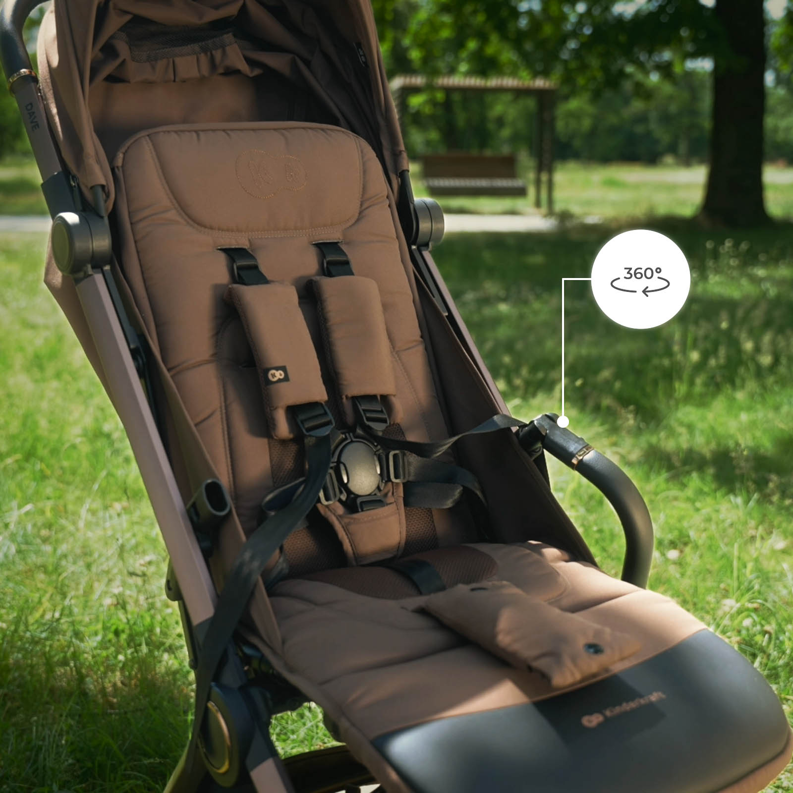 Ein brauner Kinderwagen steht im Park, mit einem 360-Grad-Symbol am Griff.