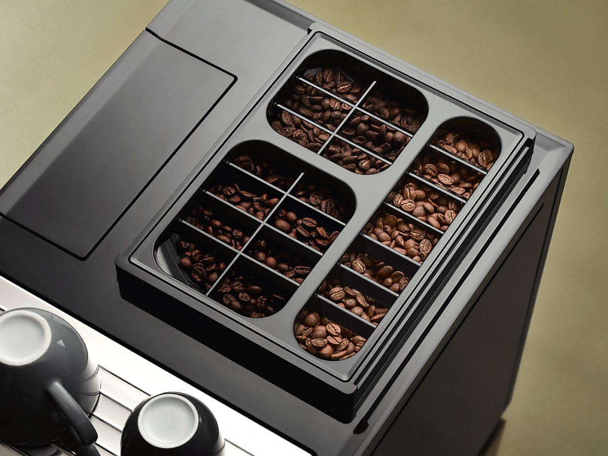 Draufsicht auf eine Kaffeemaschine mit drei Fächern voller Kaffeebohnen und zwei Tassen im Vordergrund.