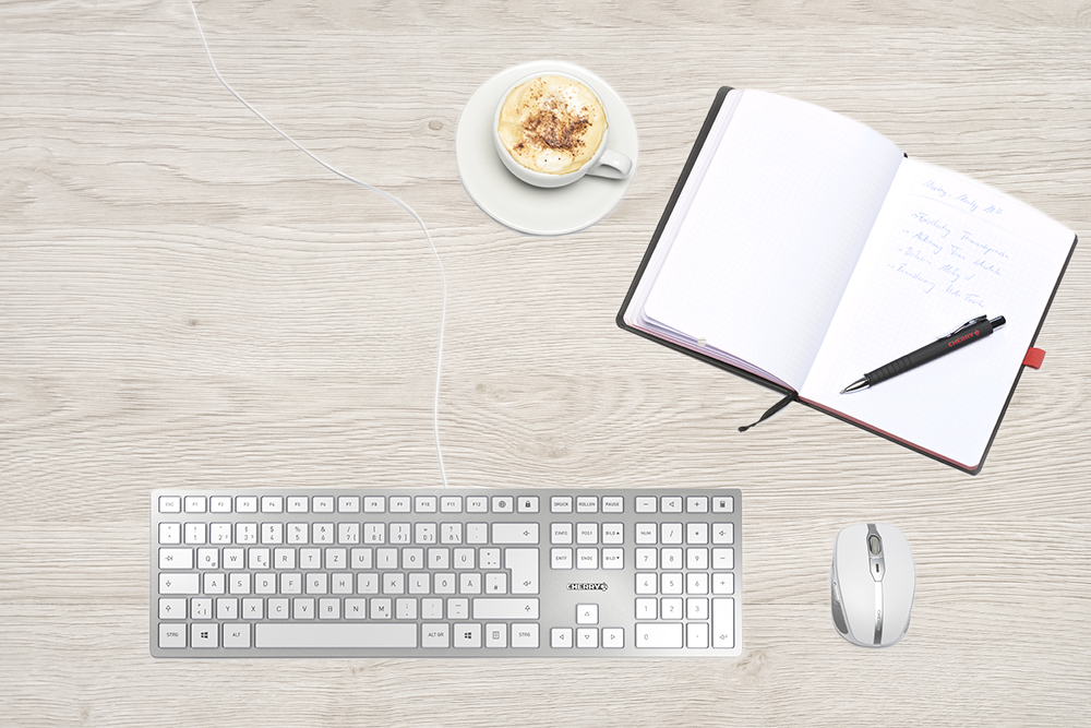 Ein Holztisch mit einer weißen Tastatur, Maus, Kaffeetasse und einem offenen Notizbuch mit Stift.
