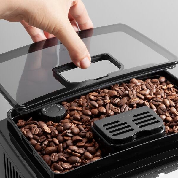 Una mano apre il coperchio scuro di una macchina da caffè piena di chicchi marroni.