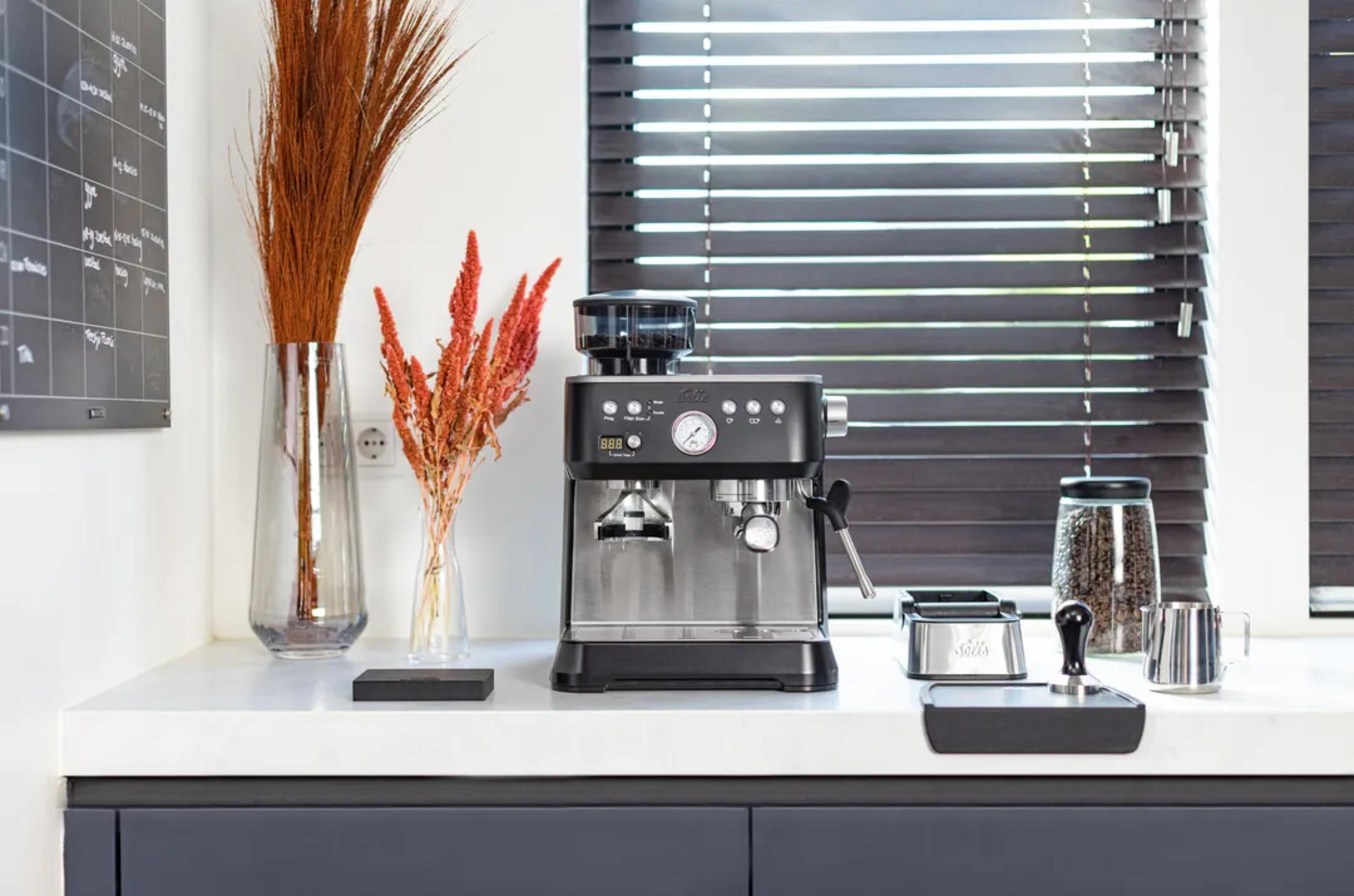 Una macchina da caffè nera su un piano cucina con vasi di fiori e un barattolo di caffè davanti a una finestra.