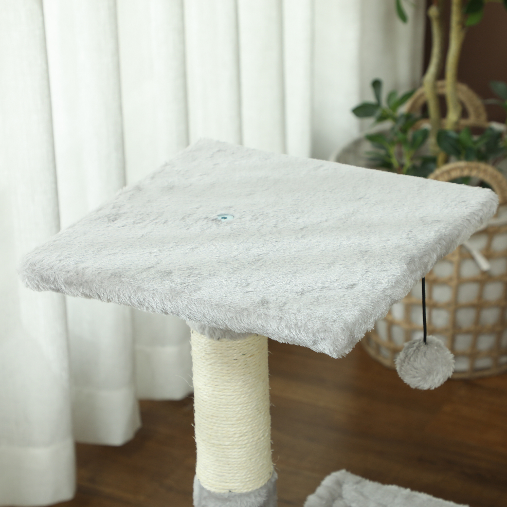 Un rascador gris para gatos con plataforma de felpa, poste de sisal y bola colgante ante una cortina.