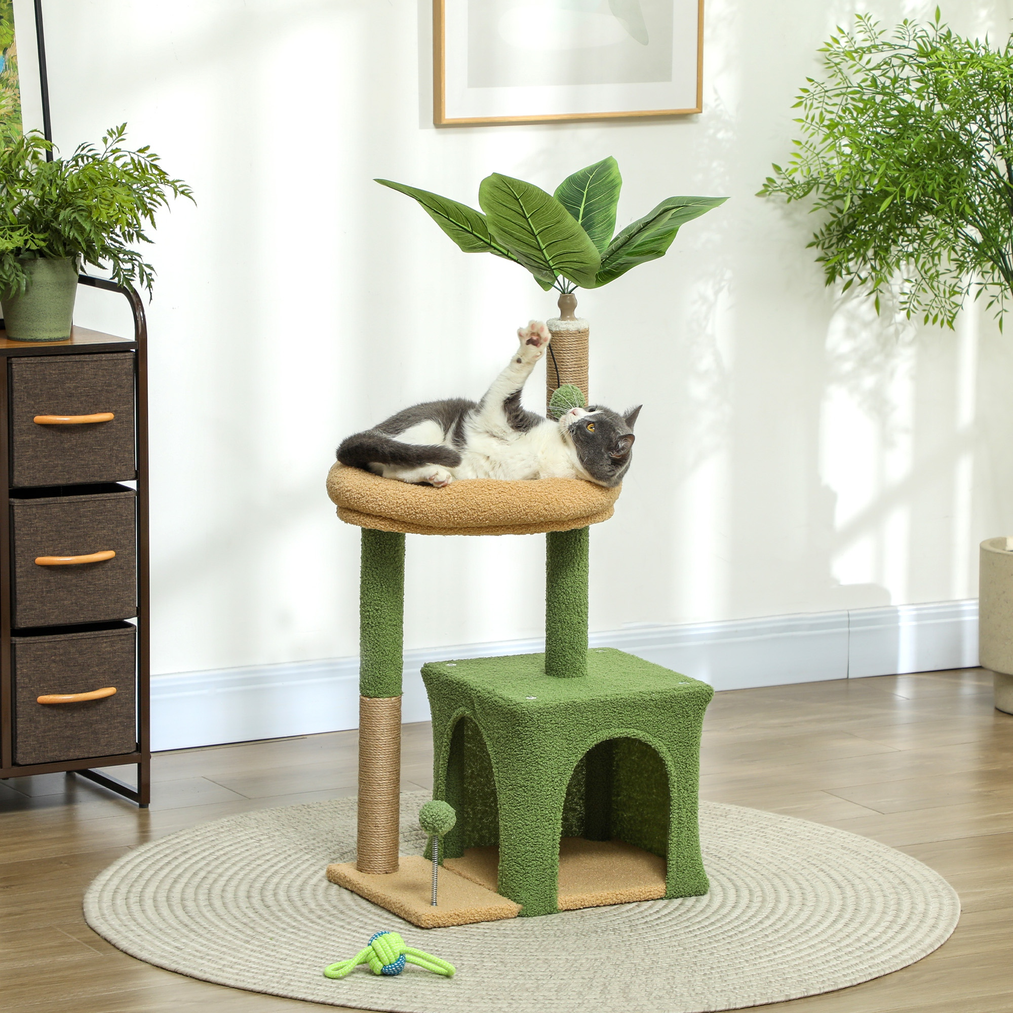 Un gato gris y blanco yace en un árbol para gatos verde con una casa y hojas artificiales.
