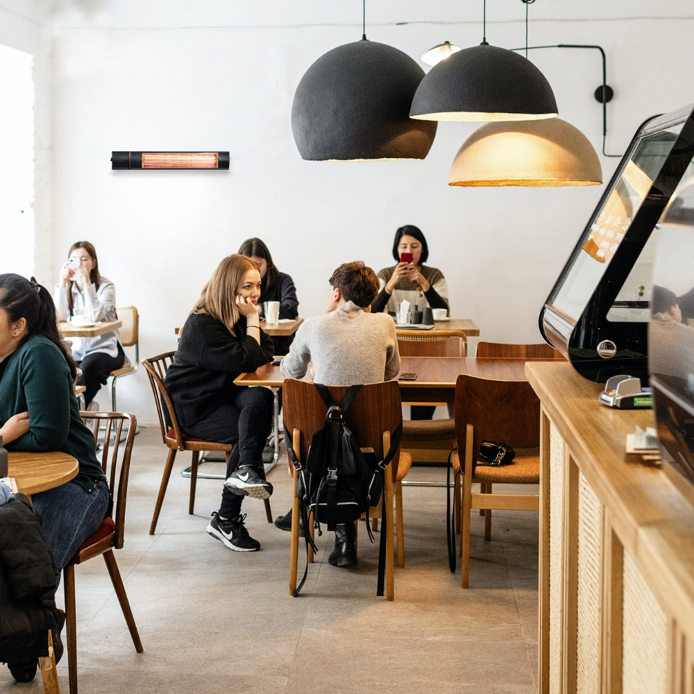 Personas se sientan en mesas de madera en un café moderno bajo grandes lámparas colgantes y un calefactor.