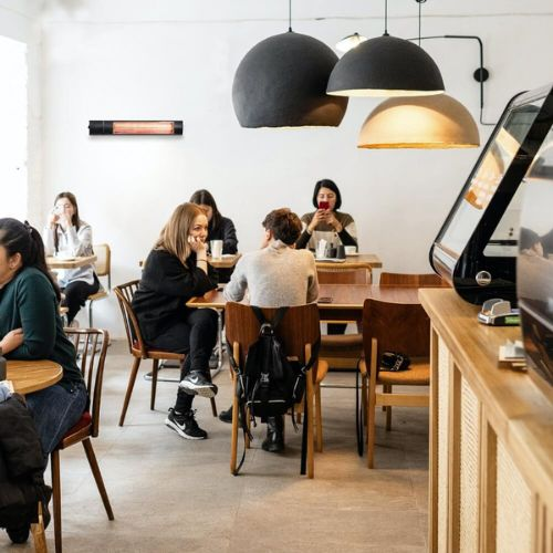 Personas sentadas en mesas en una cafetería luminosa con lámparas grandes y oscuras y un calefactor en la pared.