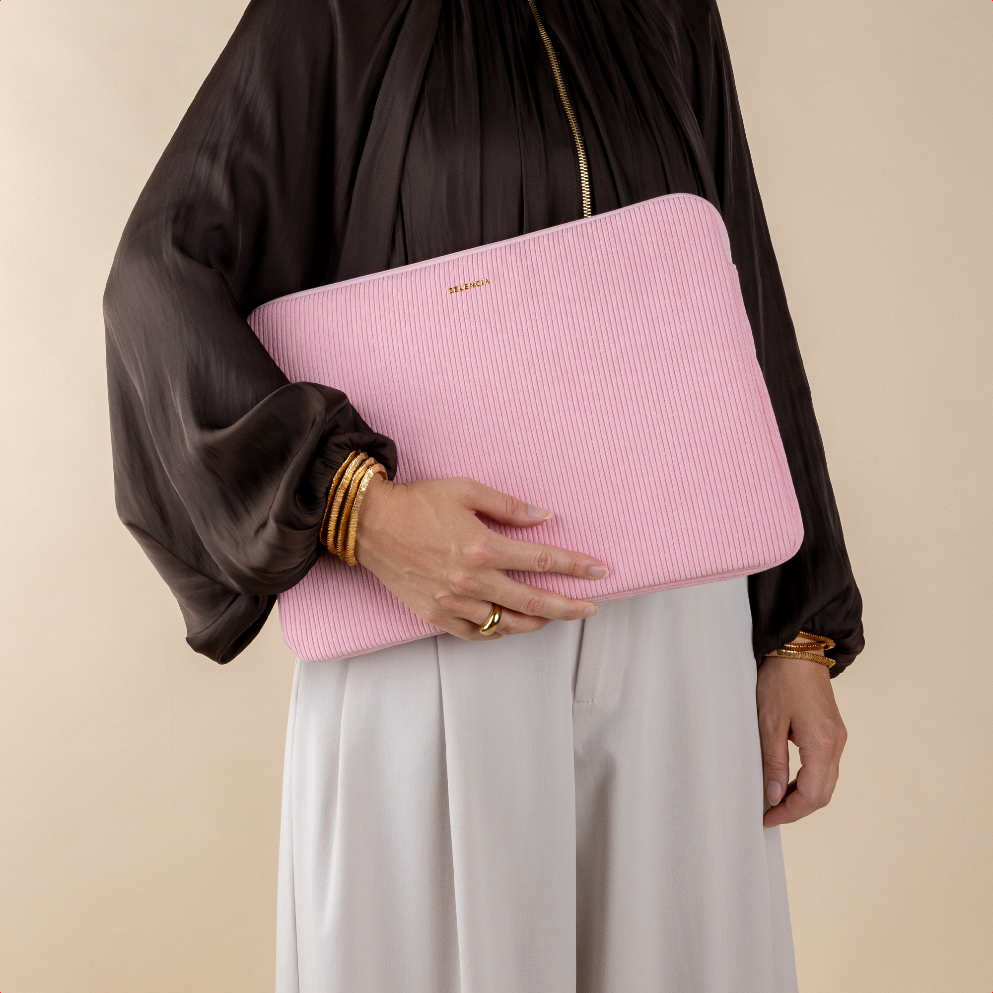 Eine Person im schwarzen Oberteil und hellen Hosen hält eine rosa gerippte Laptoptasche mit goldenen Armbändern.