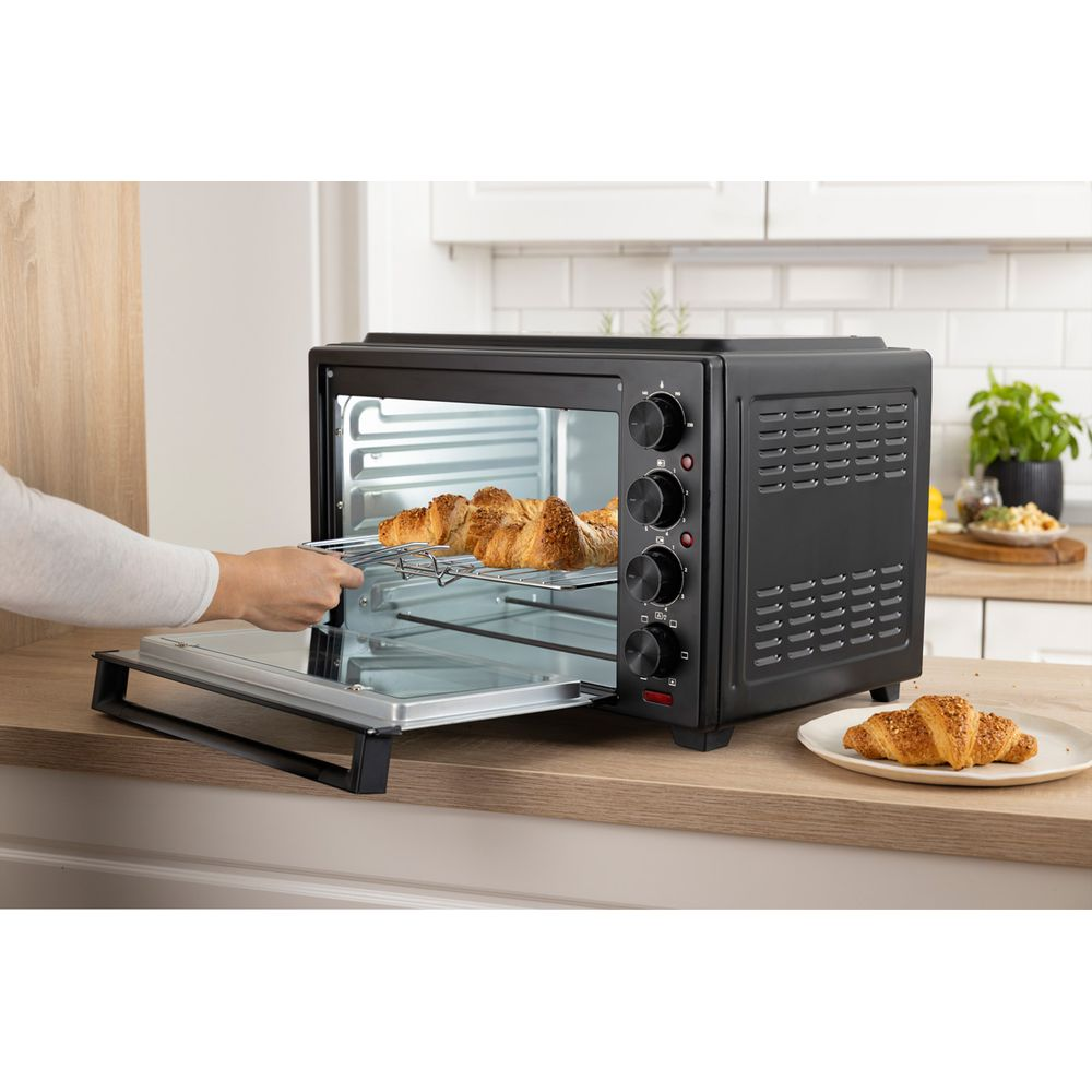 Una mano infila una griglia con croissant in un mini forno nero aperto su un piano cucina.