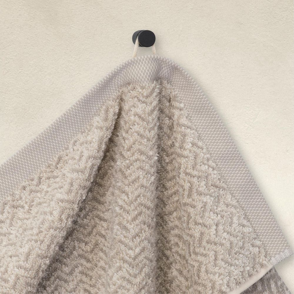 Ein beige Handtuch mit Fischgrätenmuster hängt an einem schwarzen Haken an einer hellen Wand.