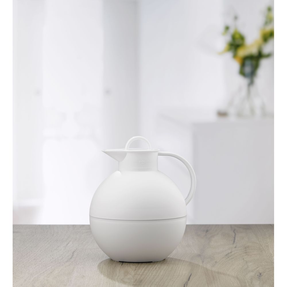 Eine weiße, kugelförmige Thermoskanne steht auf einem Holztisch, im Hintergrund ist ein helles Zimmer mit Blumen.