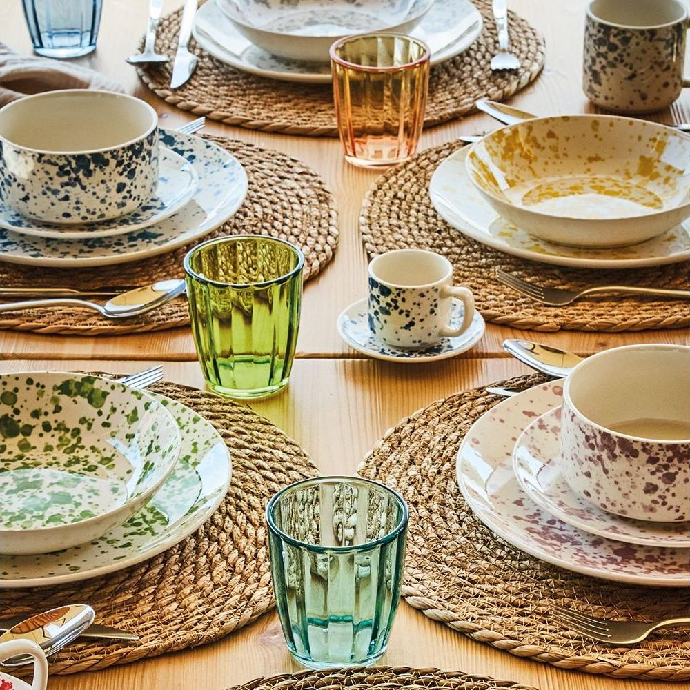 Ein hölzerner Tisch mit vier gedeckten Plätzen, bunten Tellern, Schalen und Gläsern auf Bast-Platzsets.