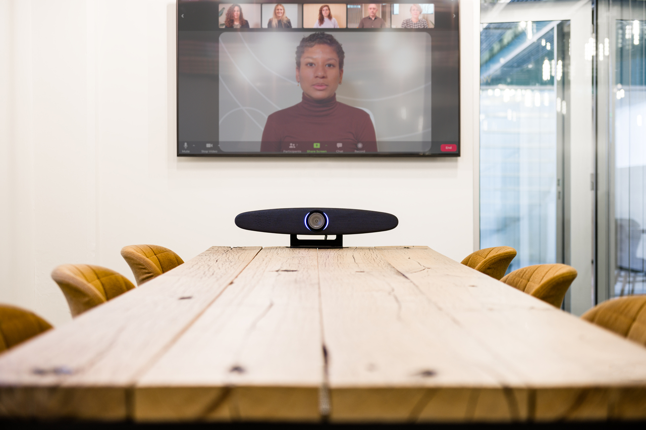 Ein Konferenzraum mit Holztisch, gelben Stühlen und einem Videokonferenzbildschirm an der Wand.