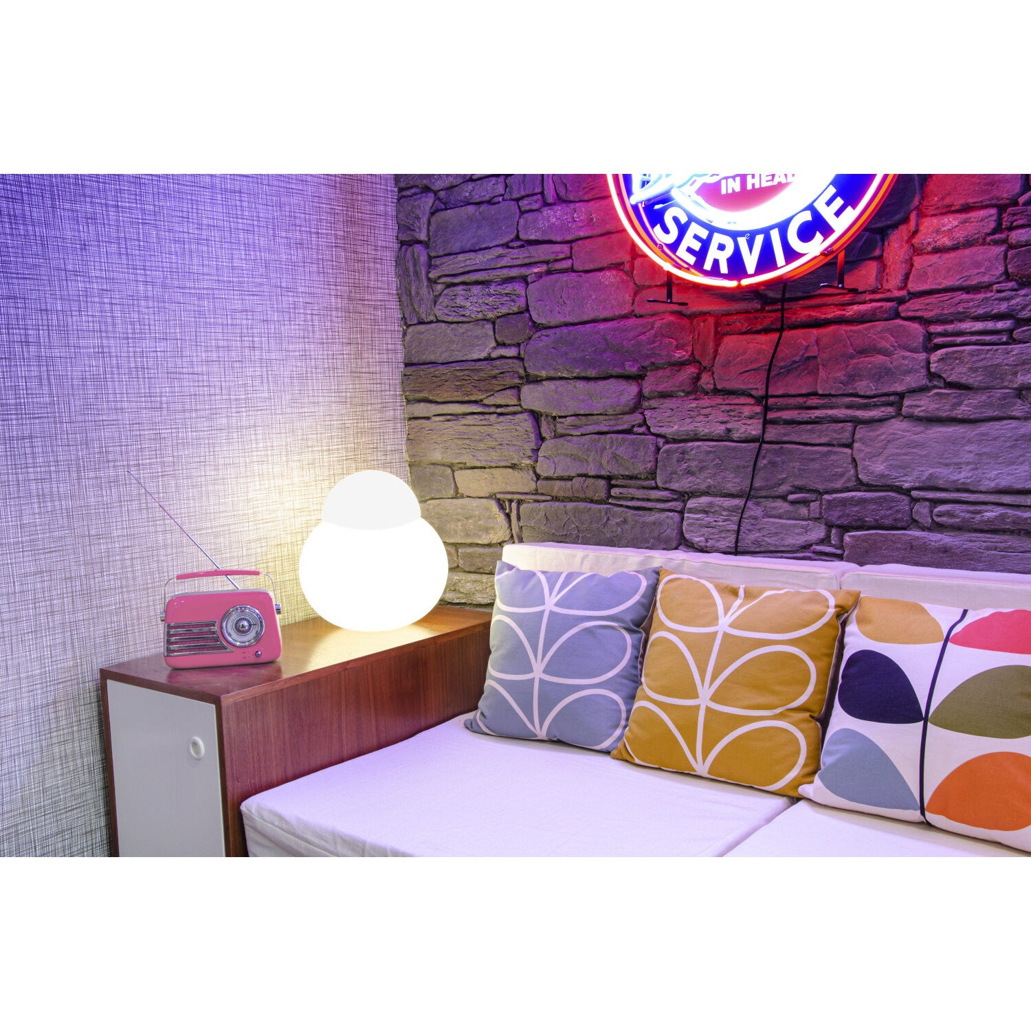 Una lampada bianca luminosa e una radio rosa sono su un mobile in legno accanto a un divano con cuscini fantasia.