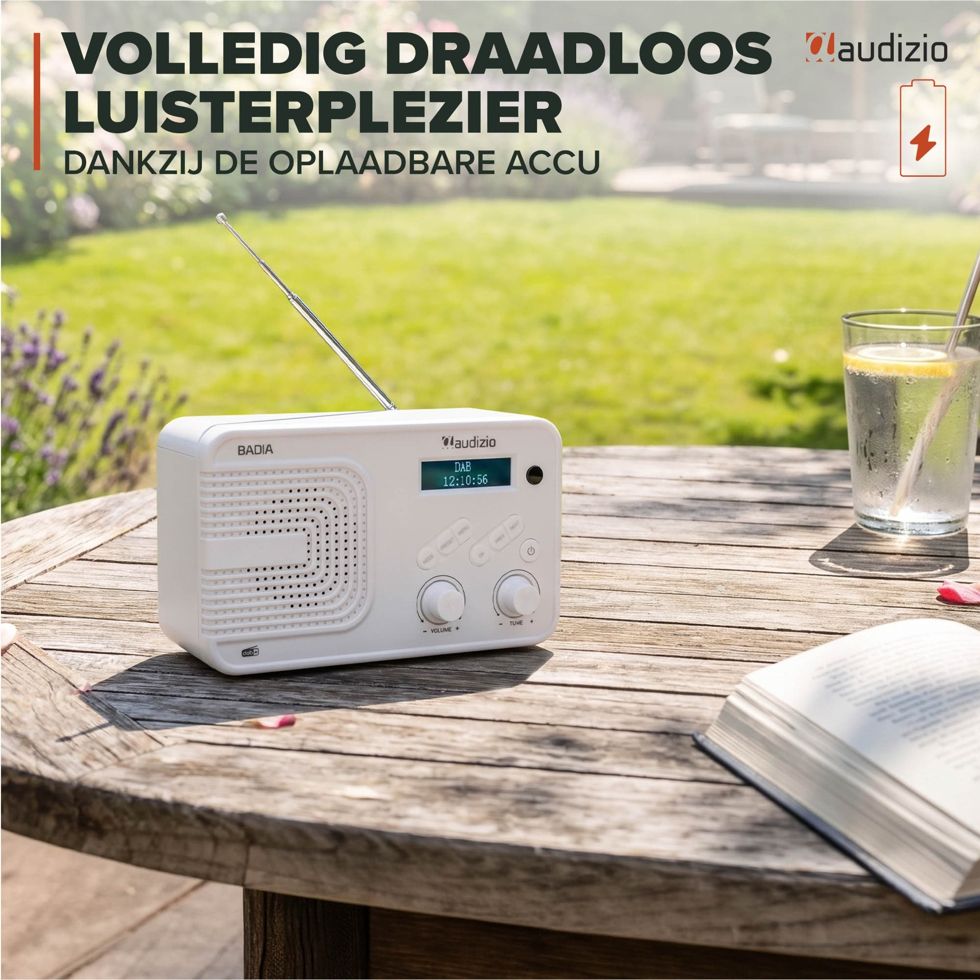 Radio Audizio blanca, vaso de agua con limón y libro abierto en una mesa de jardín de madera sobre césped verde.