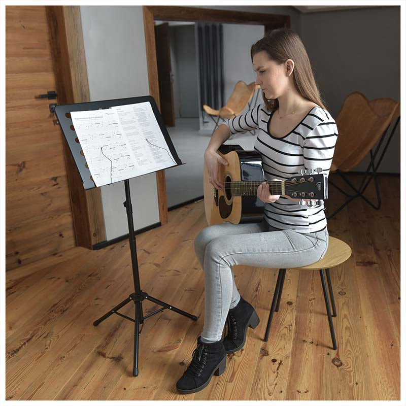 Eine Frau spielt Akustikgitarre und sitzt auf einem Hocker neben einem Notenständer.