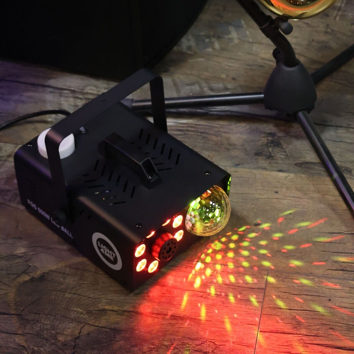 Eine schwarze Nebelmaschine mit roten LEDs und einer Discokugel wirft farbiges Licht auf einen Holzboden.