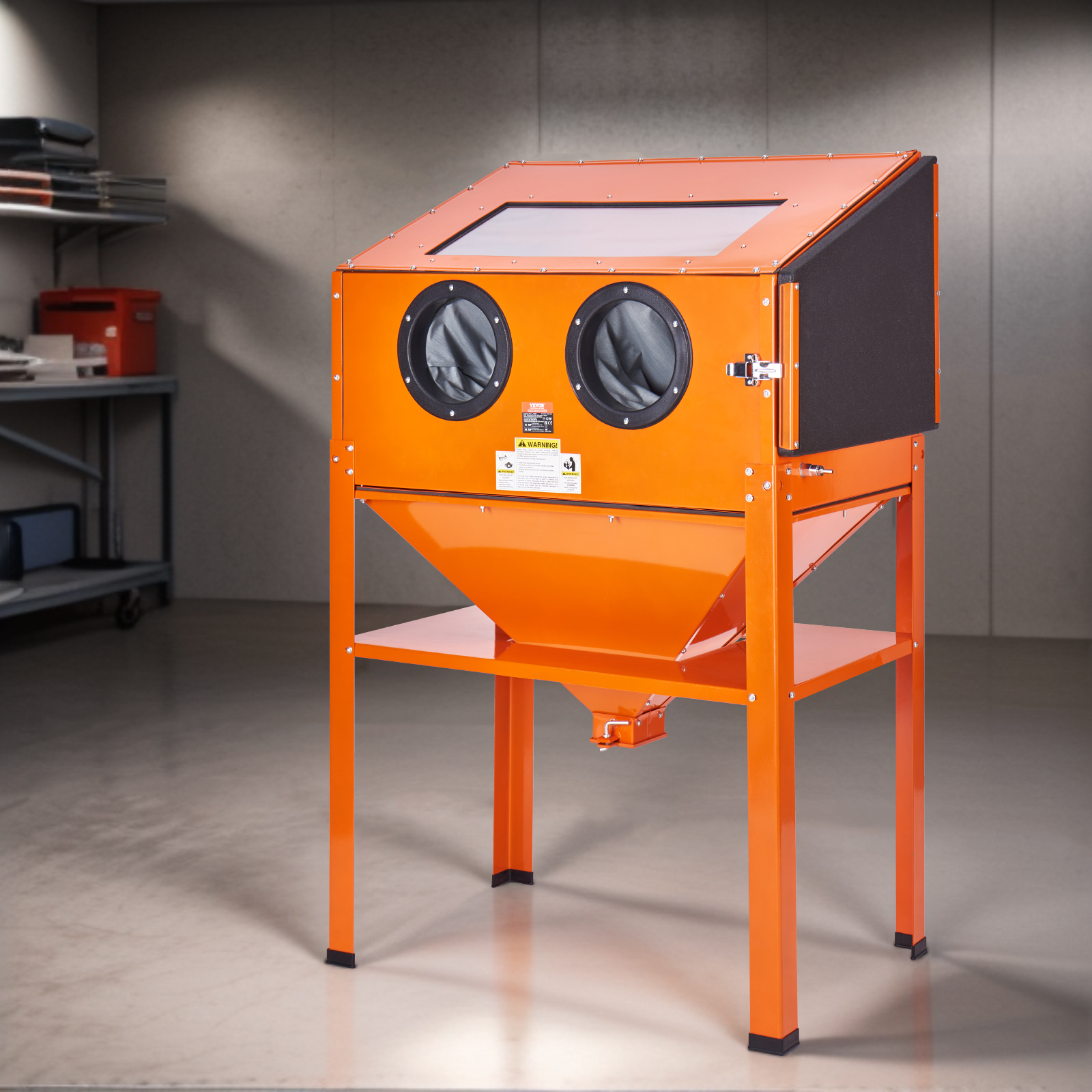 Cabina de arenado naranja con patas, dos puertos para guantes y una ventana de visualización en un taller.
