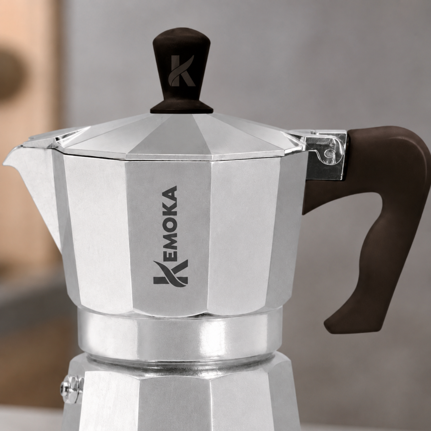 Una caffettiera moka Emoka argentata con manico e pomolo marroni su sfondo sfocato.