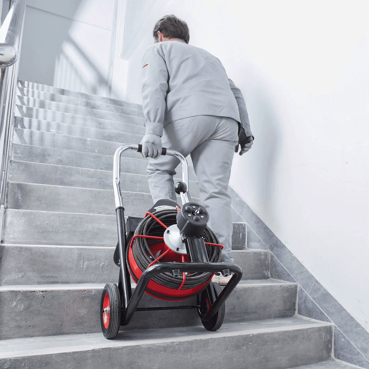 Un trabajador con ropa gris sube un carro de cables rojo y negro por una escalera.