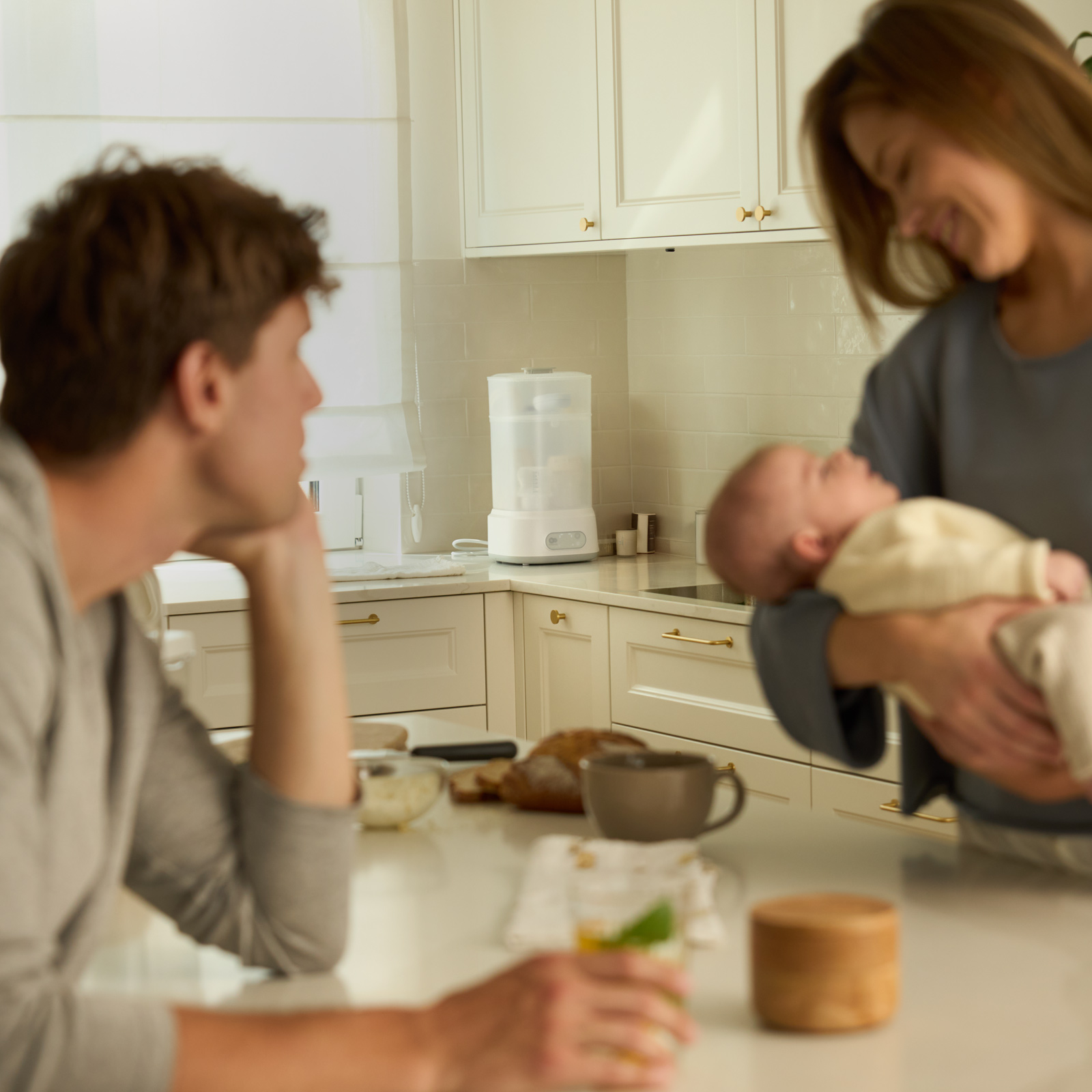 Ein Paar in einer hellen Küche, Frau hält ein Baby, Mann sitzt am Tresen.