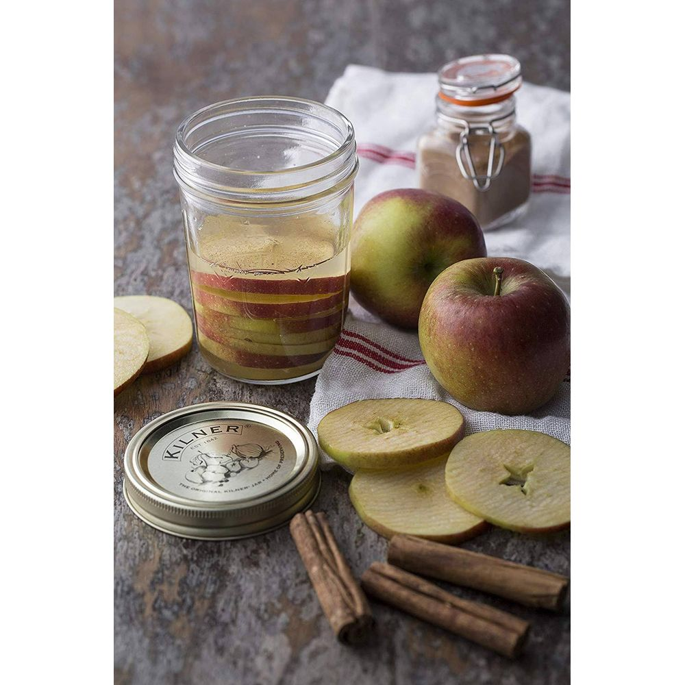 Ein Glas mit Apfelscheiben, zwei ganzen Äpfeln, Apfelringen, Zimtstangen und einem Gewürzglas auf rustikalem Holz.
