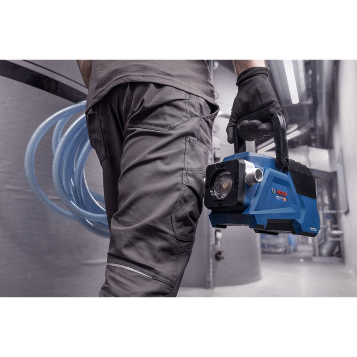Ein Arbeiter in grauer Kleidung trägt eine blaue Bosch-Pumpe und rollt einen blauen Schlauch in einem Innenraum.